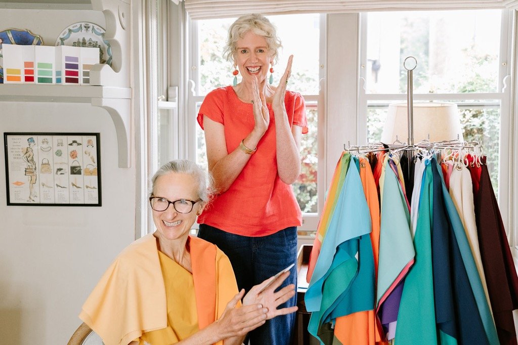 Two women, one sitting and one standing, in a room filled with colorful fabrics and a rack of various fabrics hanging, with bright natural light coming in through large windows. The woman standing is smiling widely and clapping, while the seated woman is smiling and looking at her.