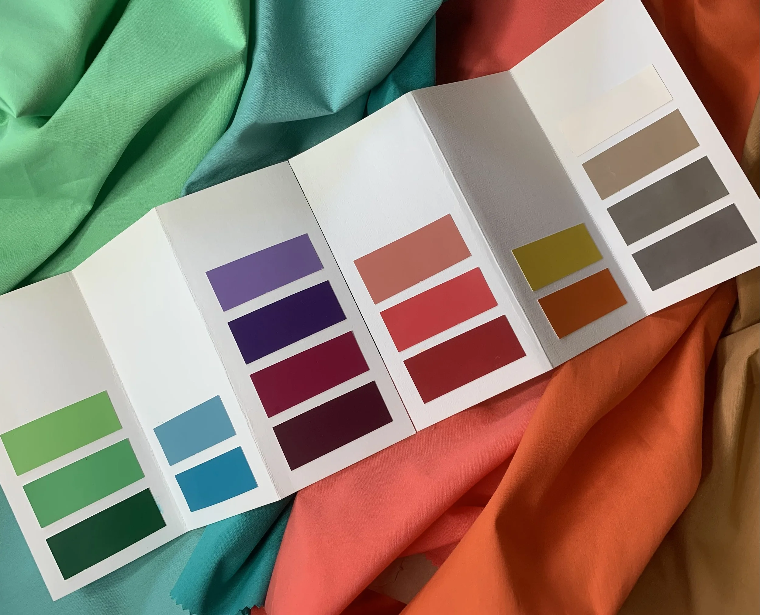 Color swatch book displaying various shades of purple, red, yellow, green, blue, brown, and gray, with fabric and paper samples in the background.