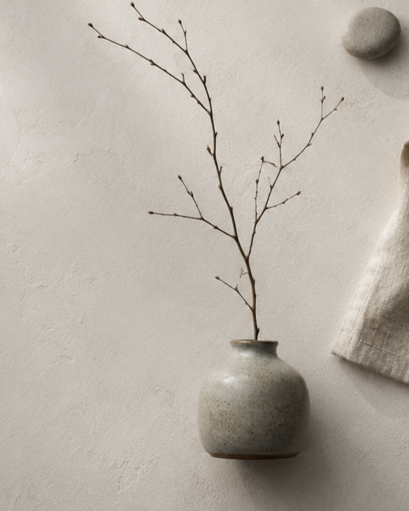 Eine Vase mit einem trockenen Zweig steht auf einer hellen Wand, daneben liegen Steine und ein Stofftuch.