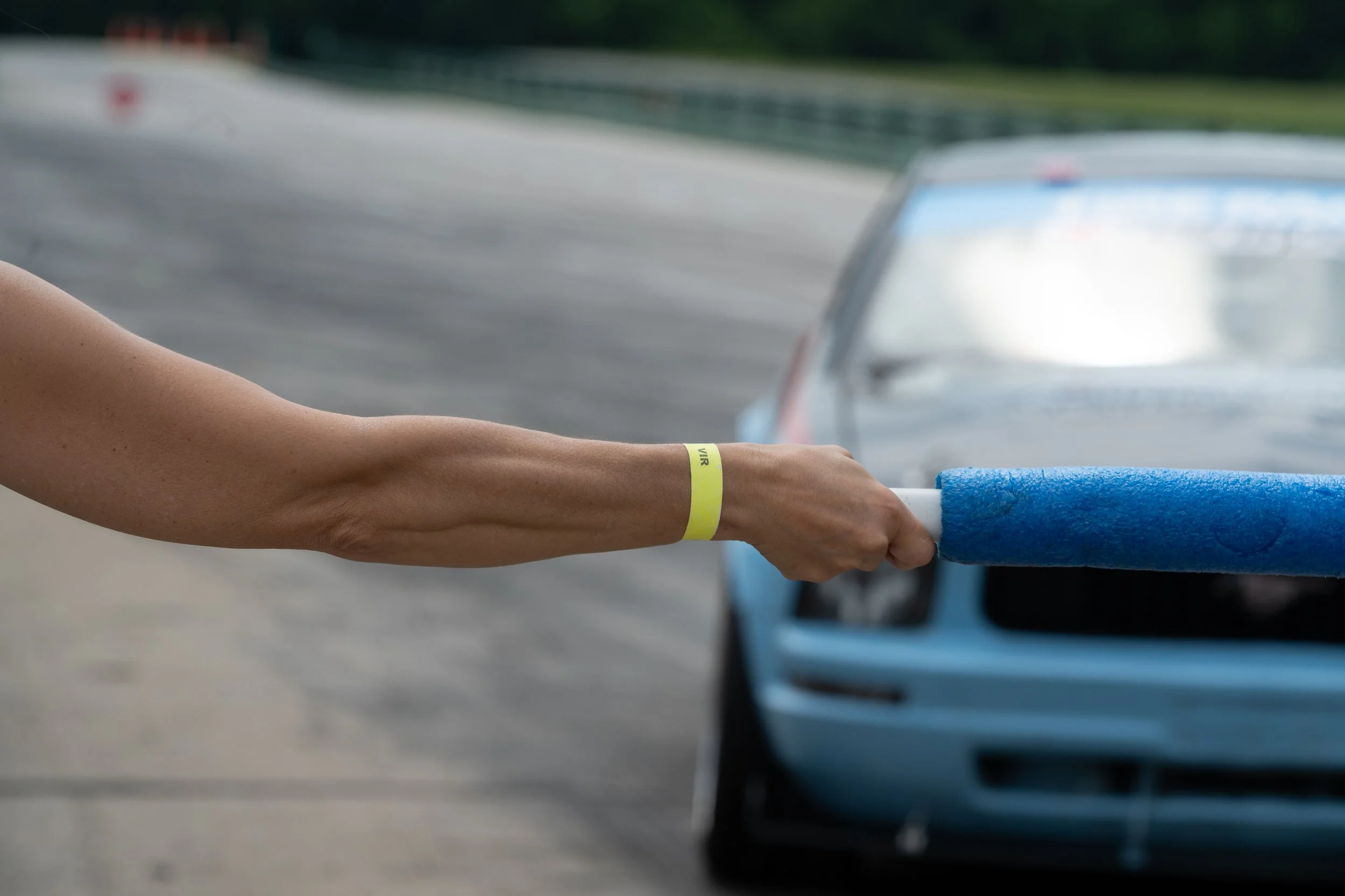 A person's arm extending from the left side of the image, wearing a yellow wristband with text, holding onto a blue foam pool noodle. In the background, there is a blurry vehicle on a road.
