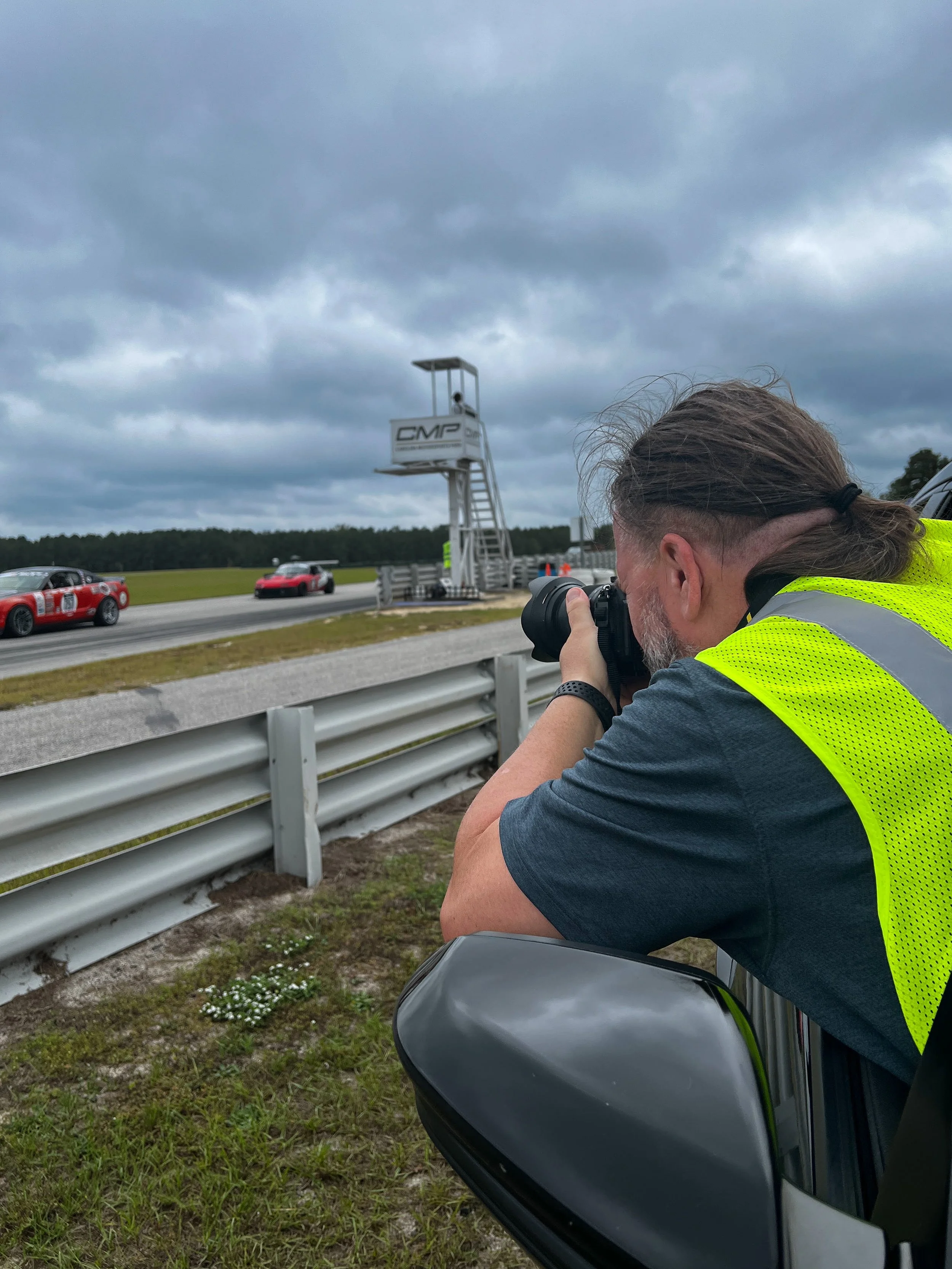 A person with long hair and a beard, wearing a high-visibility yellow vest, is taking a photograph at a motorsports event. They are squatting next to a gray motorcycle and are positioned near a metal guardrail. In the background, there are race cars on a track, a tall timing tower with a digital clock, and cloudy skies overhead.