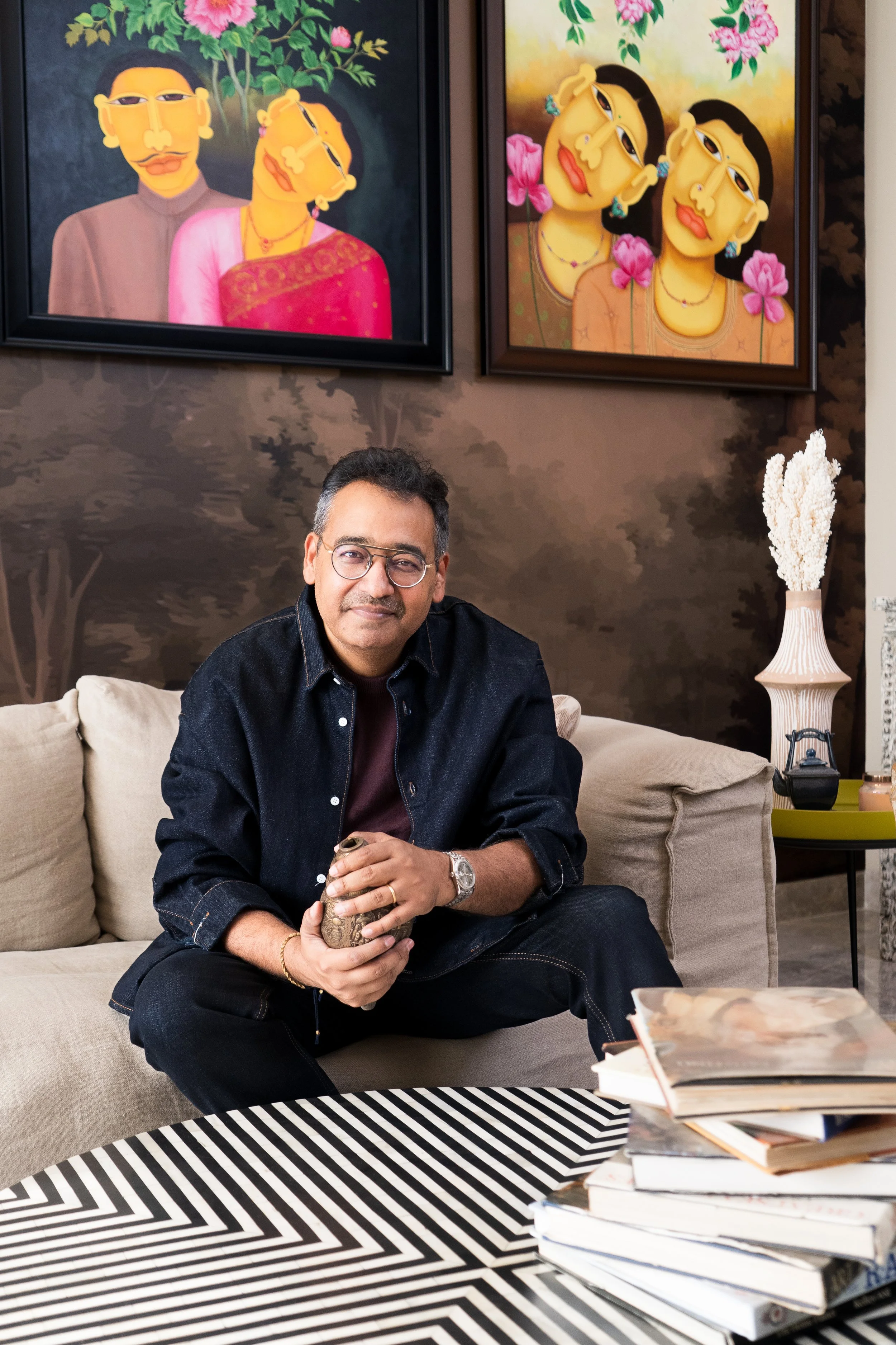 A man with glasses and dark hair sitting on a beige couch, holding a decorative object, in a room with framed artwork of stylized portraits of women hanging on the wall behind him. There are stacked books and magazines in the foreground and a vase with white decorative foliage on the side table.