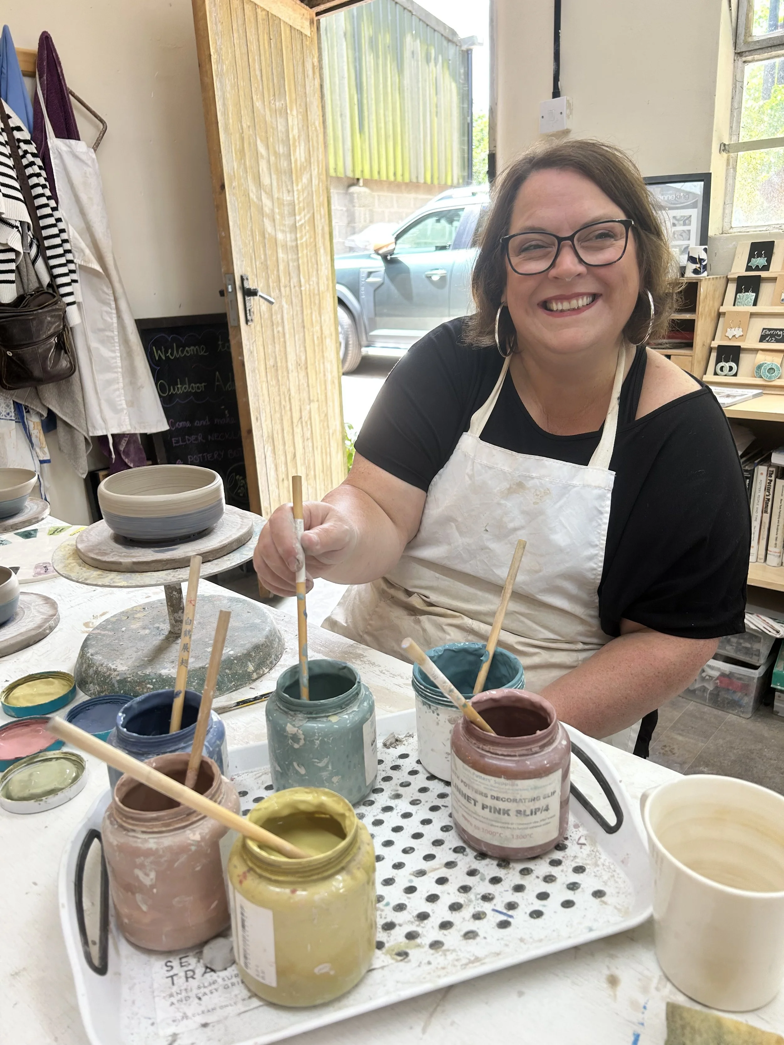 A woman with glasses and a smile, wearing a white apron, sitting at a table with various jars of colorful paint and paintbrushes for ceramics or pottery painting.