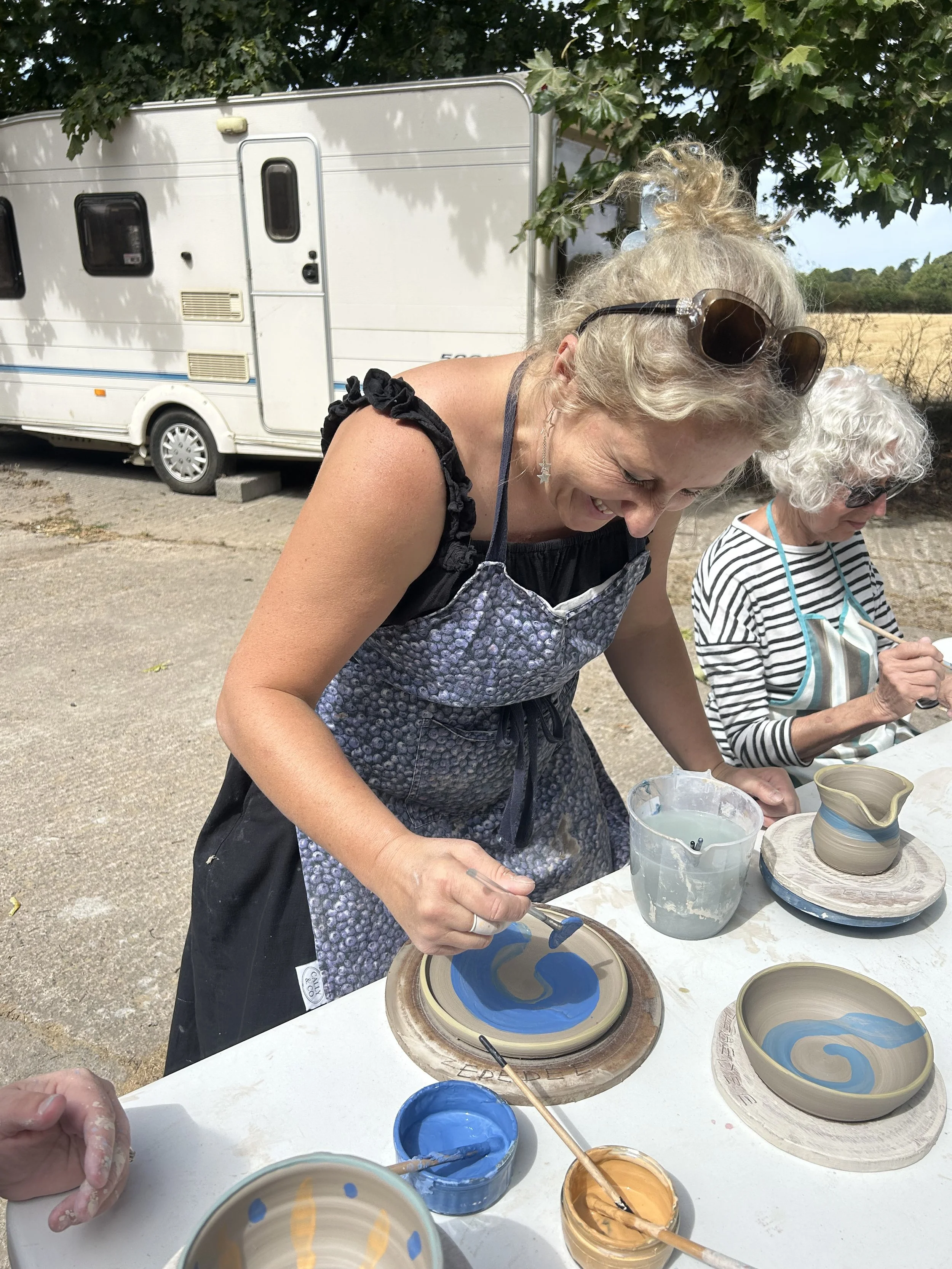 Two women are engaged in pottery painting outdoors, with a focus on the woman in the foreground who is painting a ceramic plate with blue and yellow swirls. There are several painted ceramic bowls and plates on the table, along with paint jars in blue, yellow, and orange. A motorhome is parked behind them under a tree.