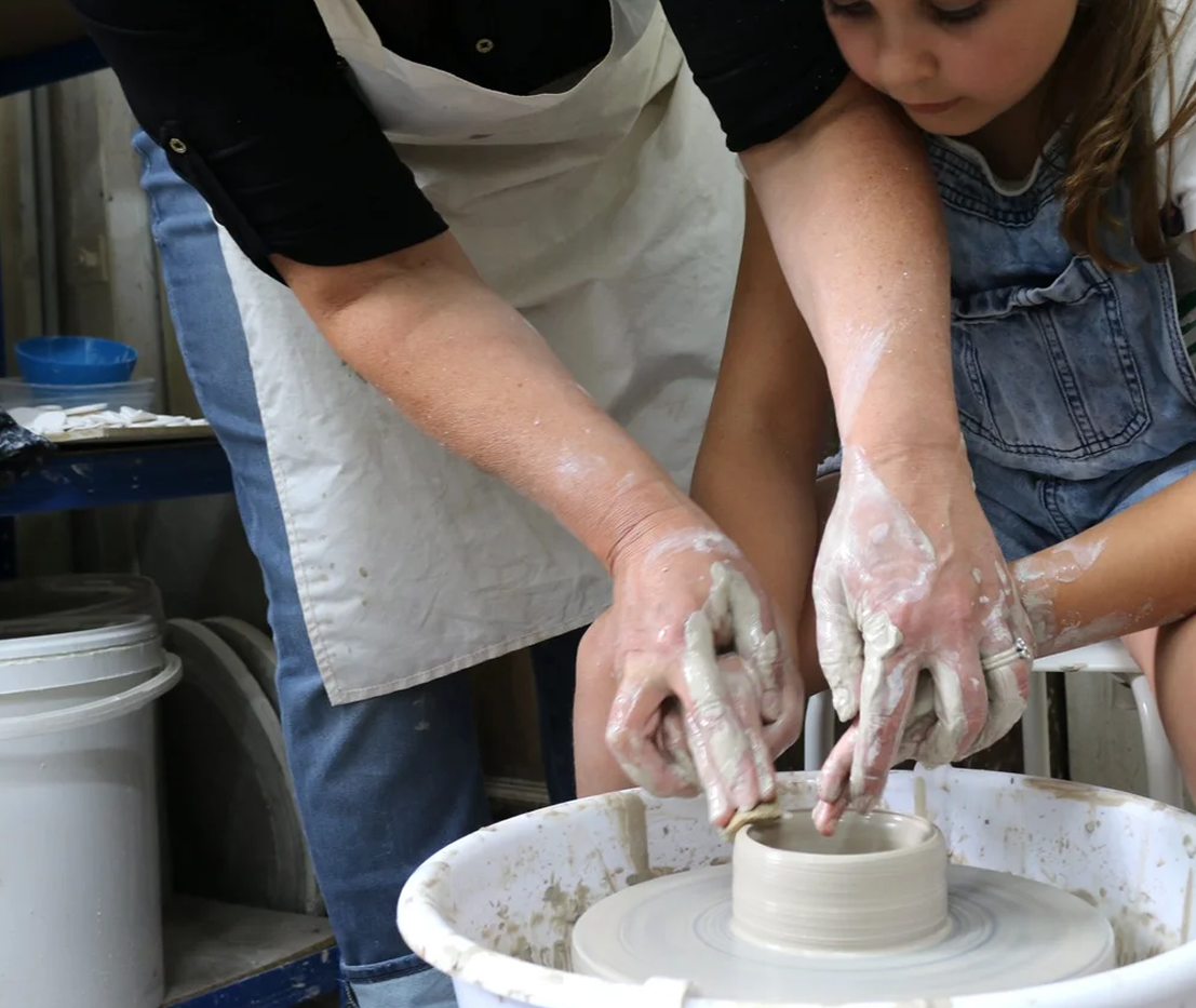 A person and a young girl are making pottery together on a pottery wheel, shaping a clay pot with their hands.