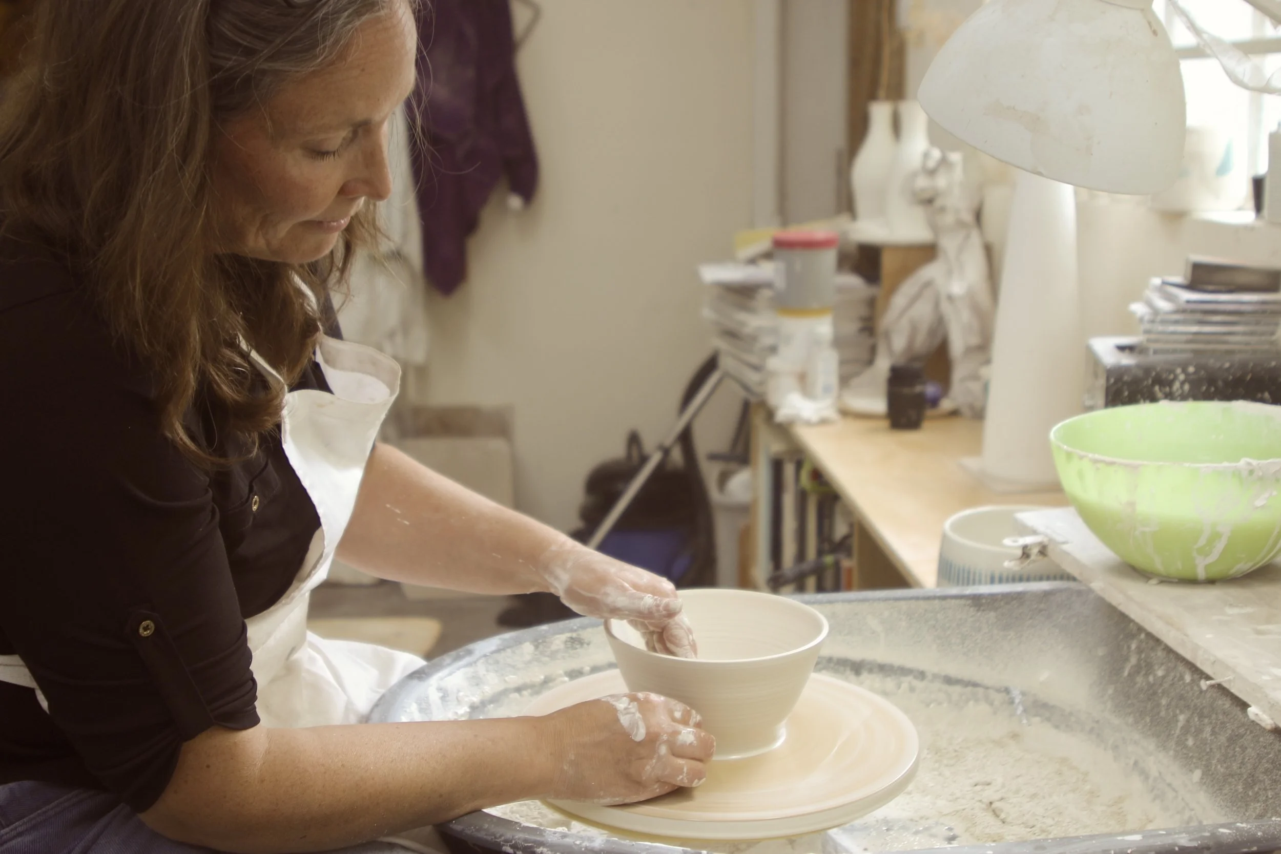 A woman working on a pottery wheel, shaping a clay bowl in a pottery studio filled with supplies and pottery equipment.