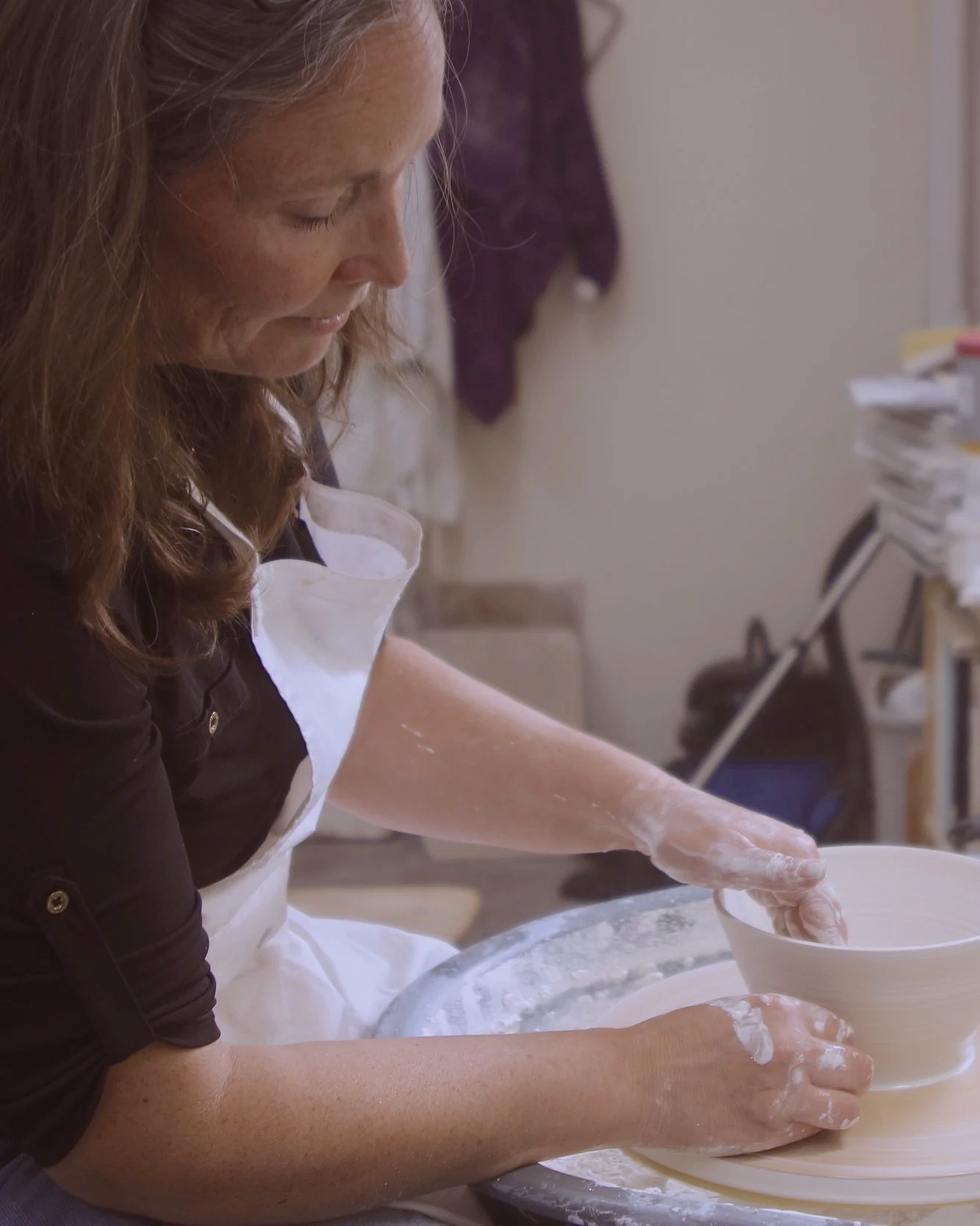 A woman wearing a white apron and black shirt is mixing dough in a white bowl at a kitchen counter.