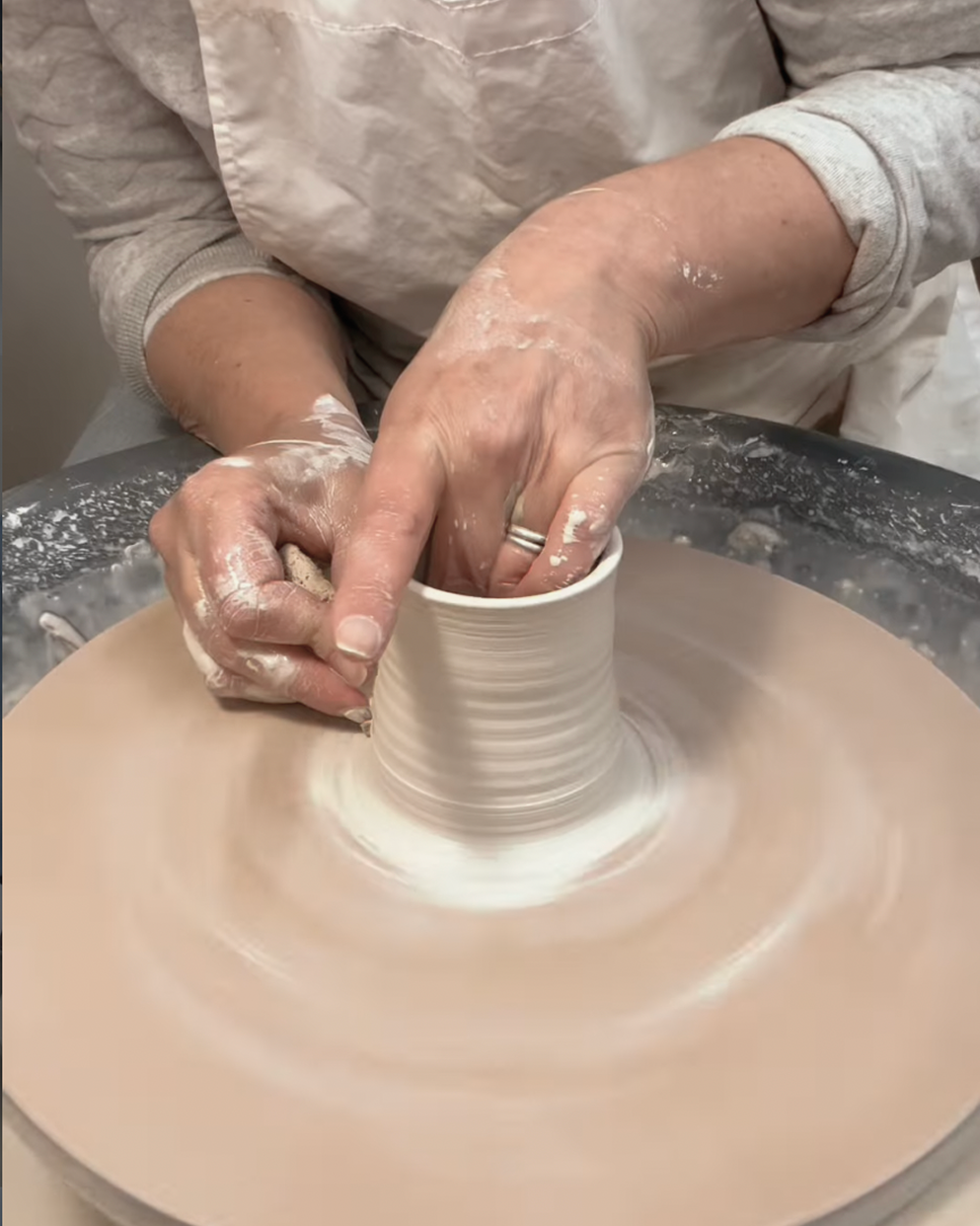 A person shaping a clay pot on a pottery wheel with their hands covered in clay.