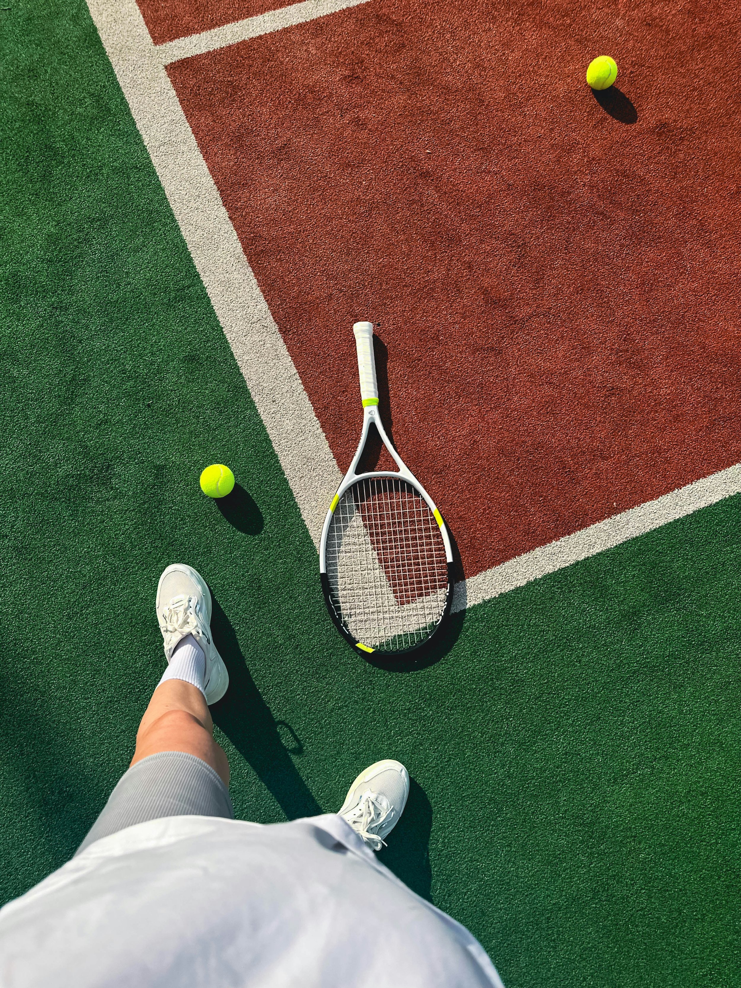 A tennis court with a tennis racket, two tennis balls, and the photographer's feet in white sneakers. The court has green and red surfaces with white lines.