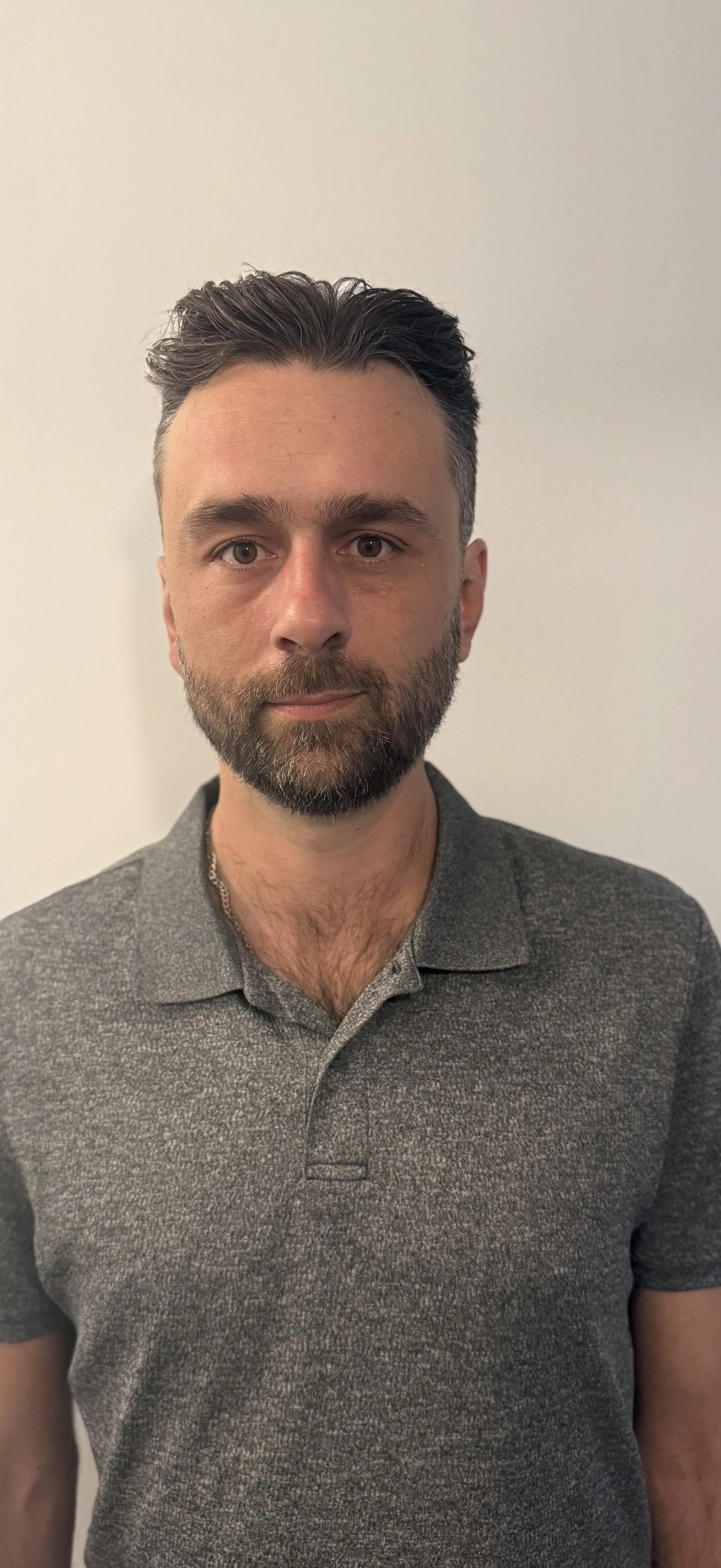 Man with dark hair and beard wearing a gray polo shirt, standing against a plain light-colored background.