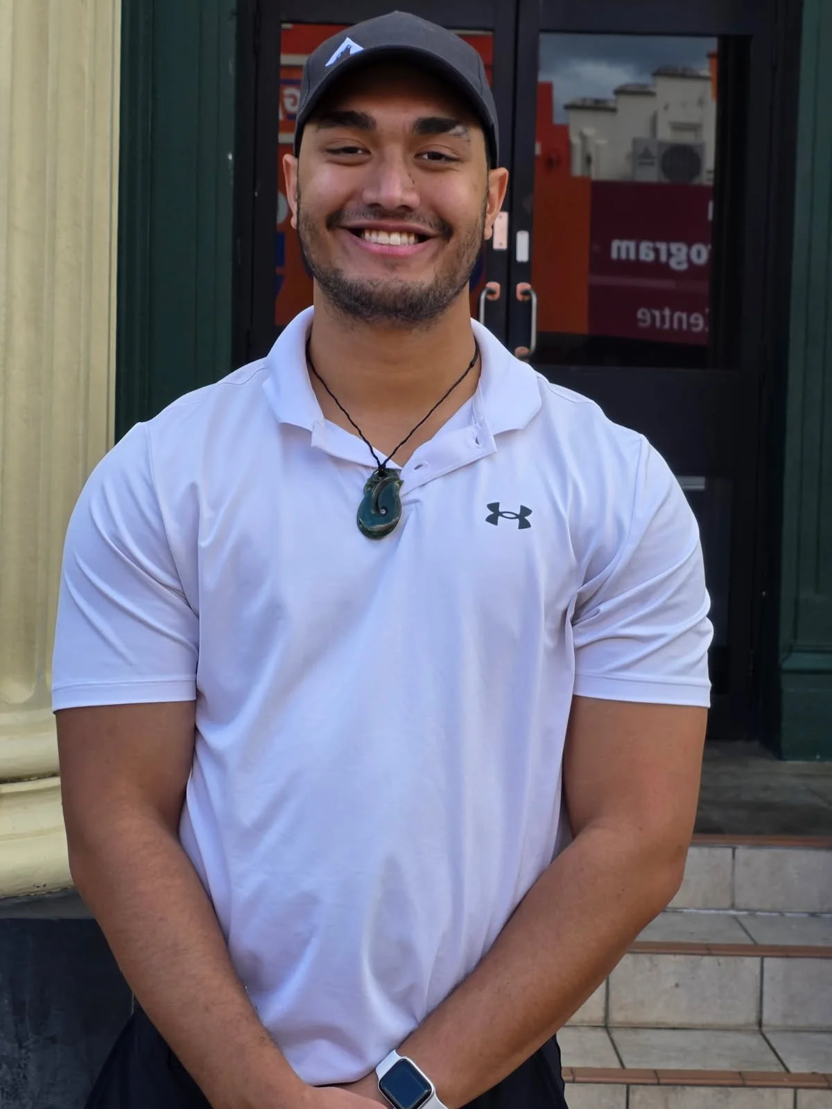 A smiling man wearing a black cap, white Under Armour polo shirt, a black necklace with a green pendant, and a smartwatch, standing in front of a glass door with a red and black sign behind him.