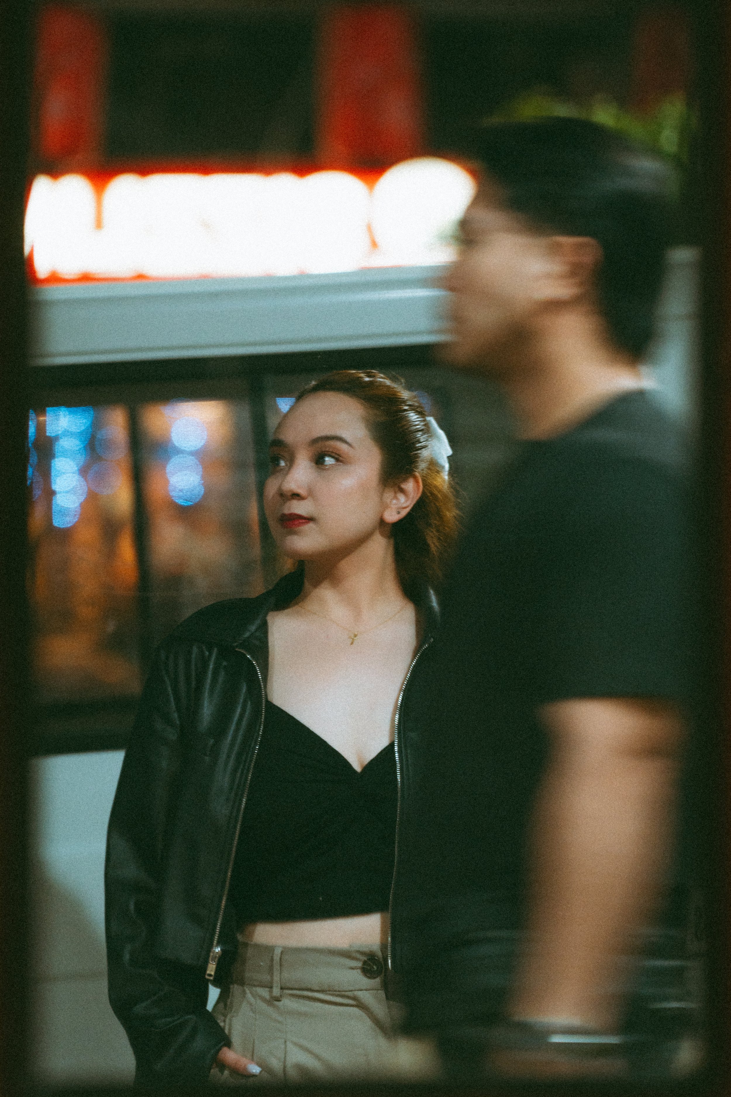 A young woman with brown hair tied back, wearing a black zip-up jacket over a black top, beige pants, and a gold necklace, looking to the side in front of reflective glass with blurred city lights in the background.