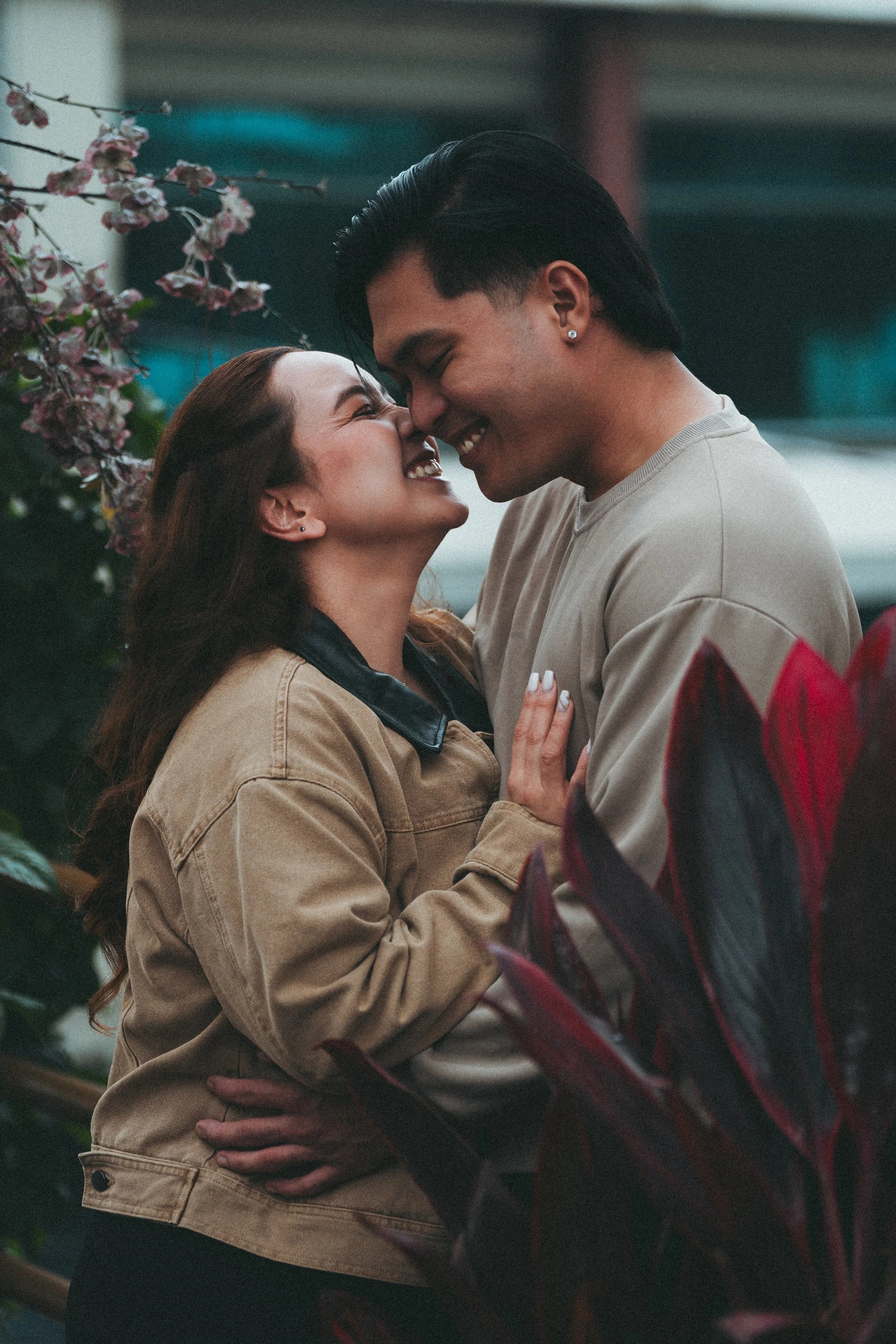 A couple smiling and touching foreheads, in a close embrace, outdoors with flowering plants around them.