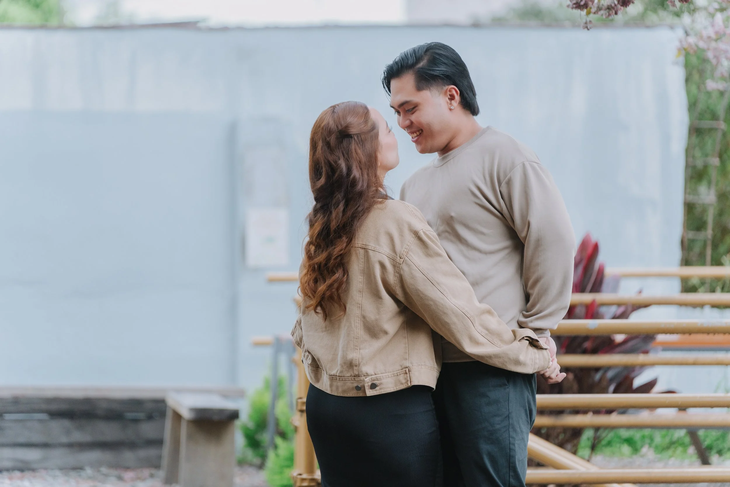 A couple smiling and embracing outdoors near a blue building with pink flowers and benches in the background.