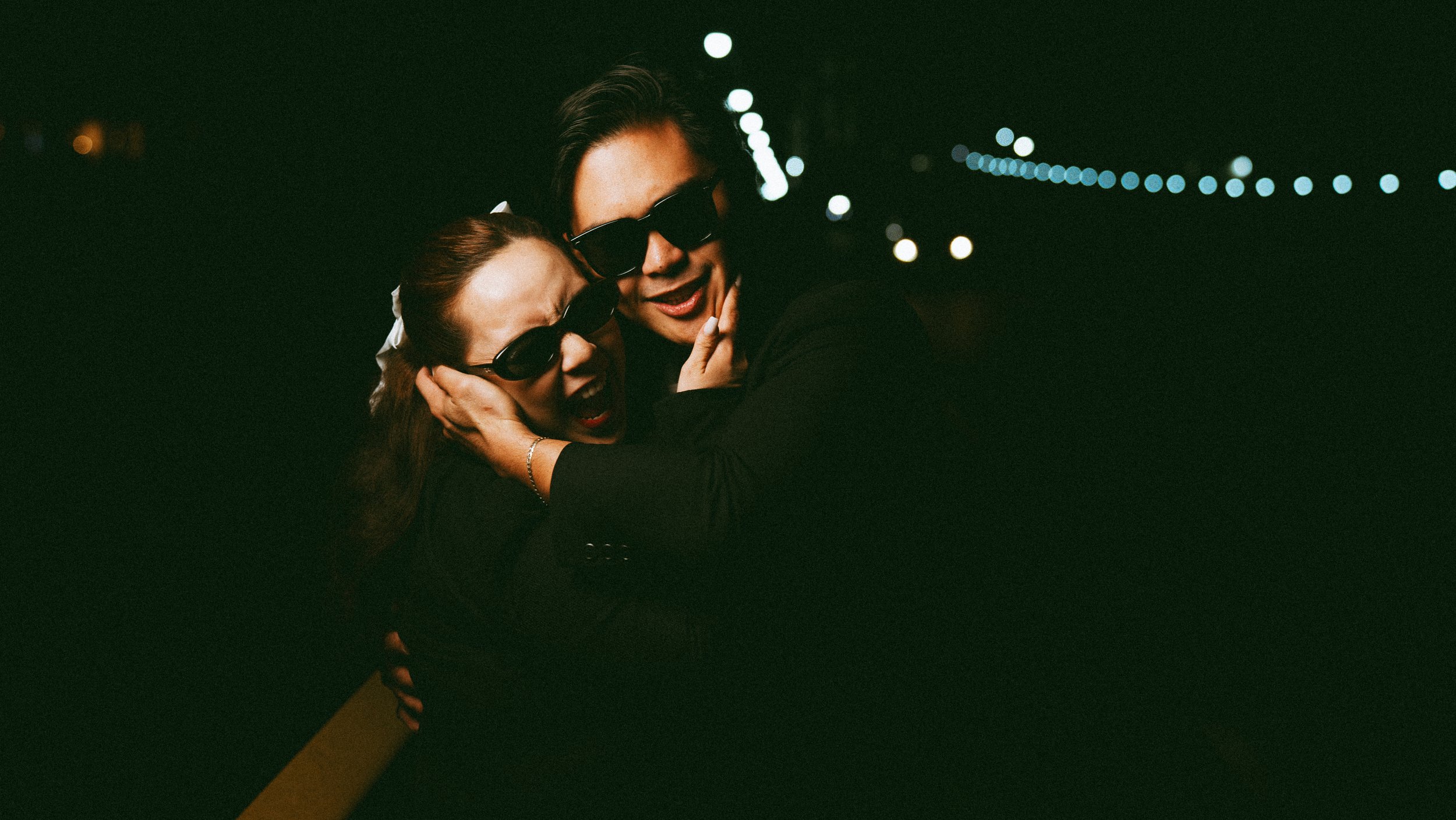 Two women with dark sunglasses hugging at night with string lights in the background.
