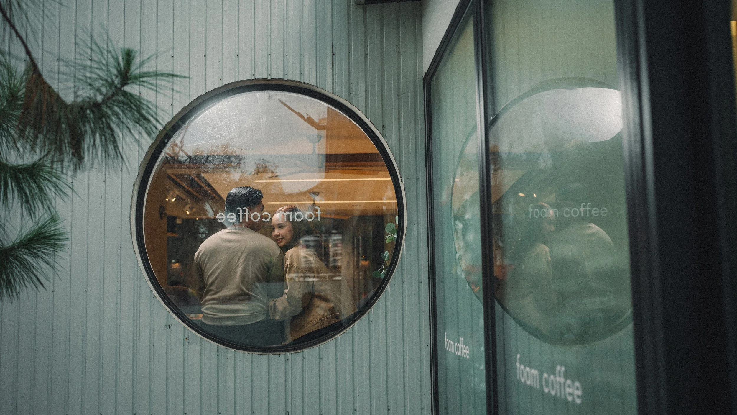 Two people are inside a coffee shop, visible through a circular window on the exterior wall of the building. They are engaging in a conversation. The exterior wall is made of vertical metal siding, and there are green trees nearby. The shop has a sign that reads "from coffee."