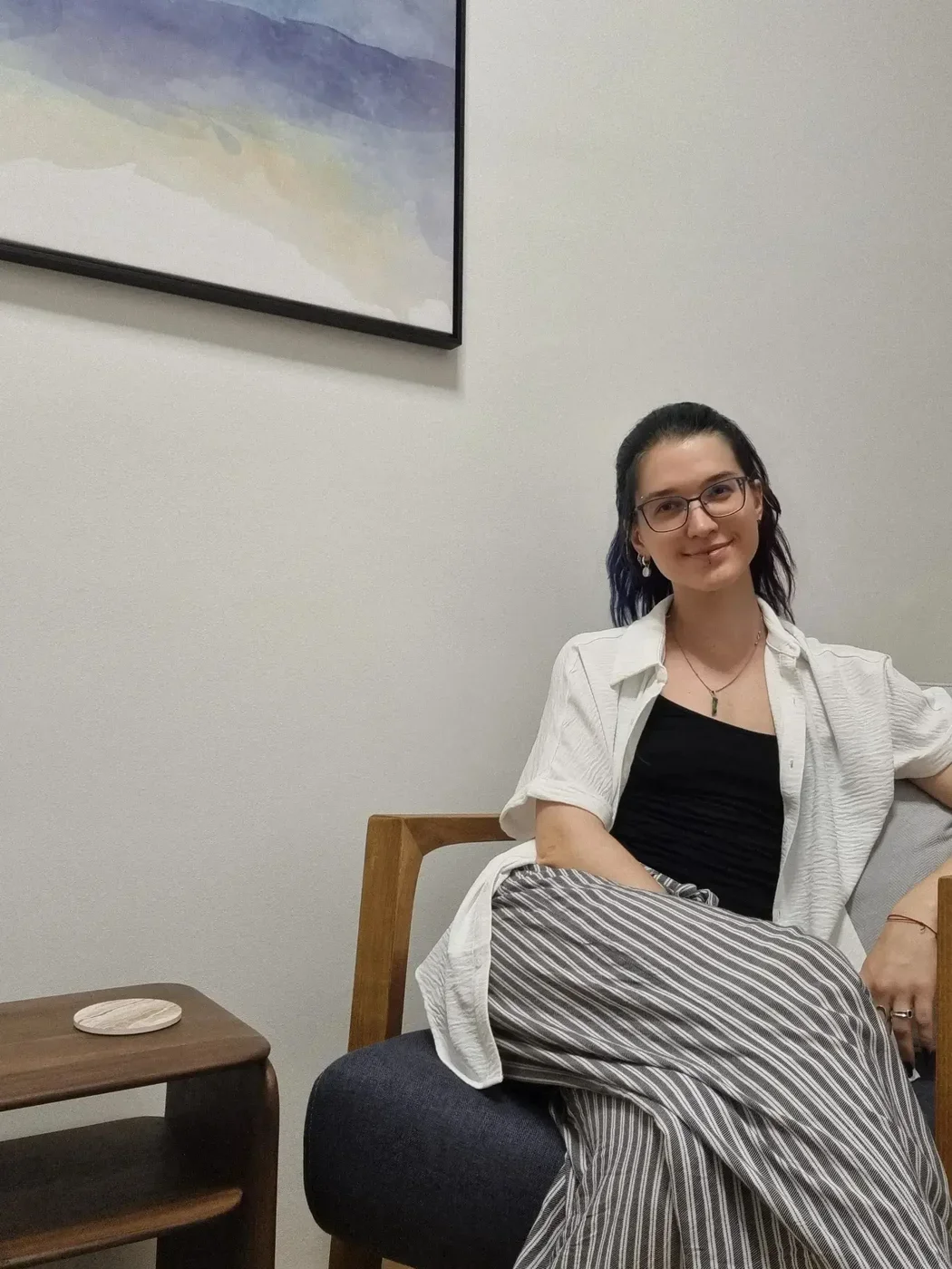 Junge Frau mit dunklen, blauen und schwarzen Haaren, Gläsern, einem weißen Hemd über einem schwarzen Top, einer Halskette und Ohrringen, sitzt auf einem schwarzen Sessel in einem Raum mit weißen Wänden und einem gemalten Bild an der Wand.