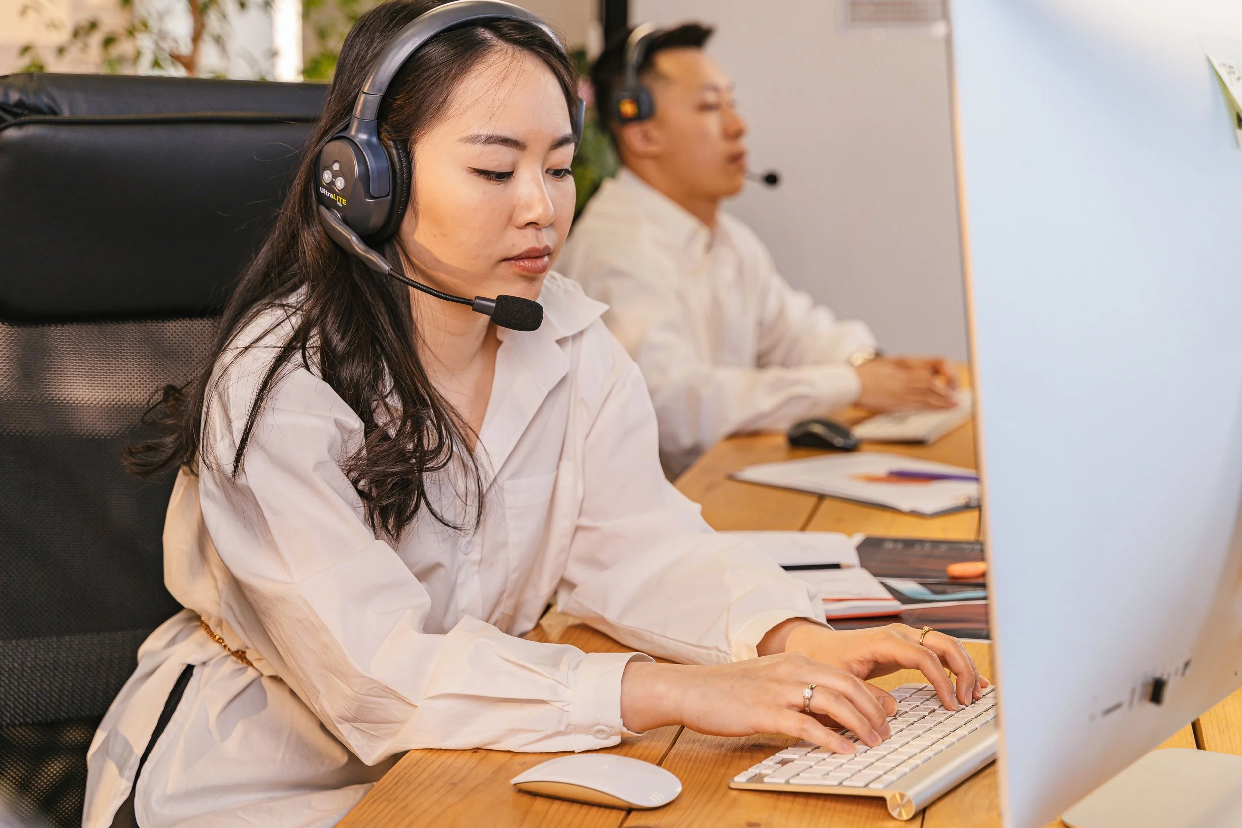Close-up of smiling woman wearing a headset with microphone, line of similar women in background, all wearing headsets, in a call center or customer service environment.