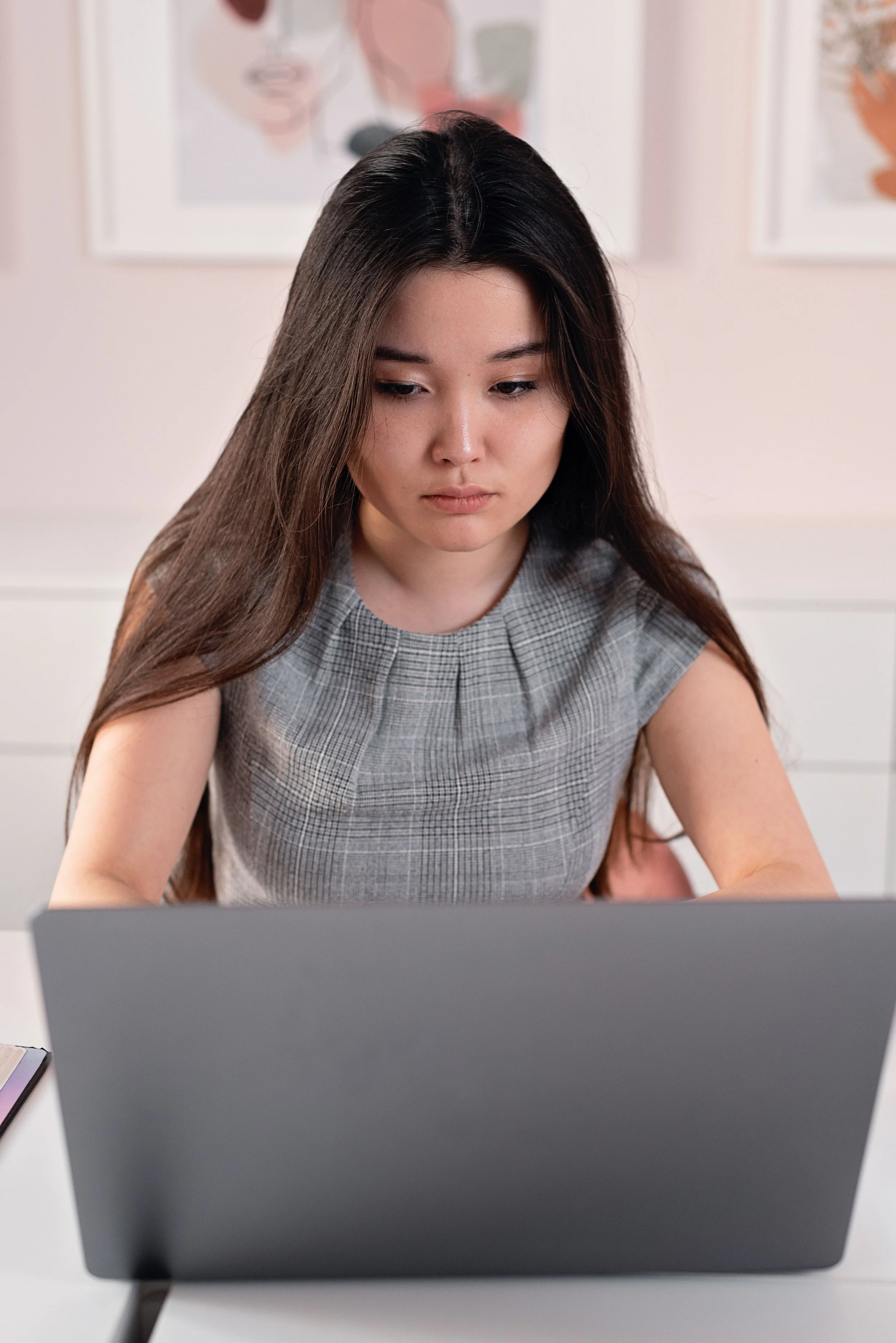 A woman with long dark hair working at a desk, looking at a laptop screen, with framed abstract artwork on the wall behind her.