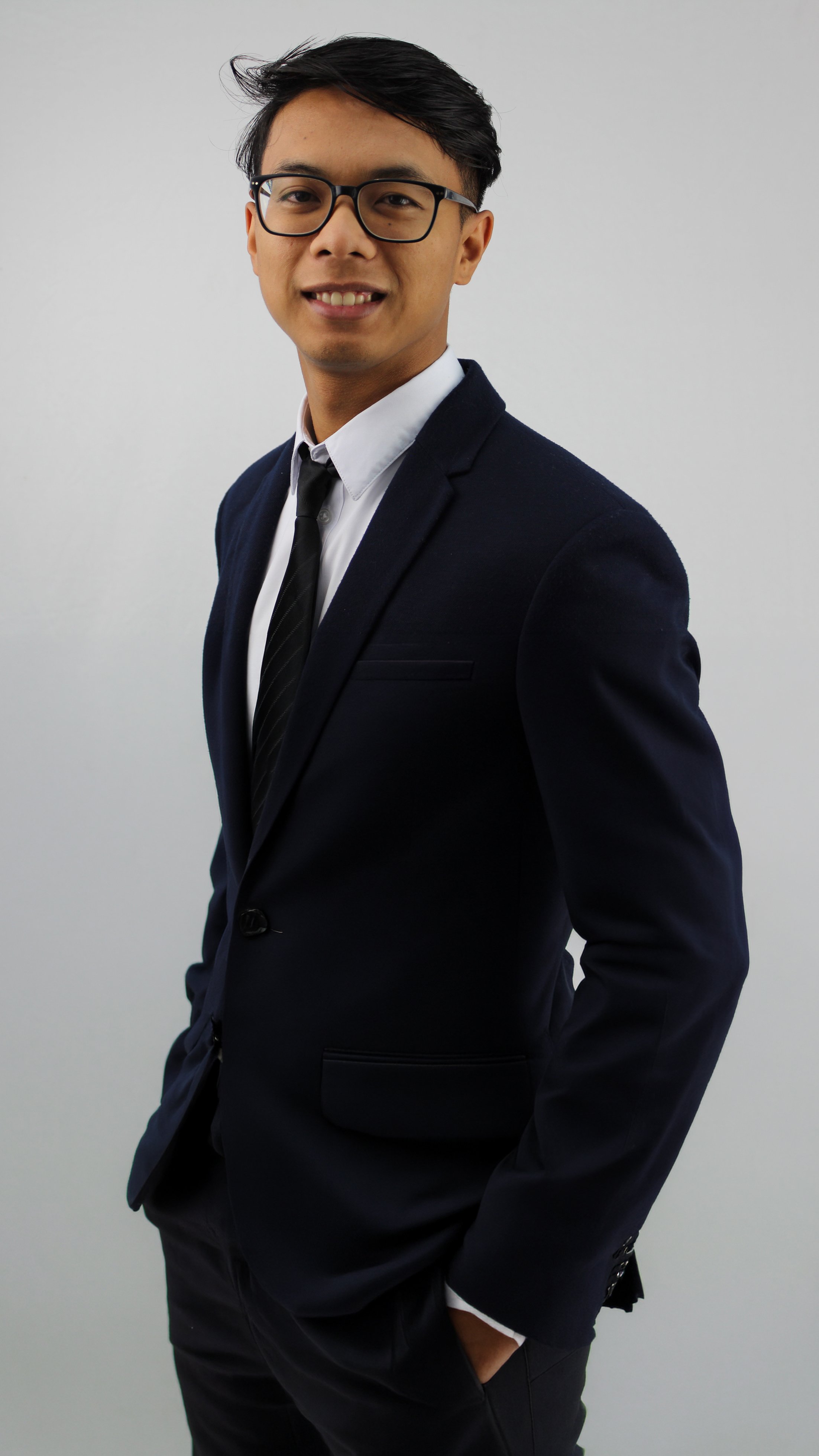 A young Asian man in a dark suit, white shirt, black tie, and glasses, smiling and posing against a plain gray background.