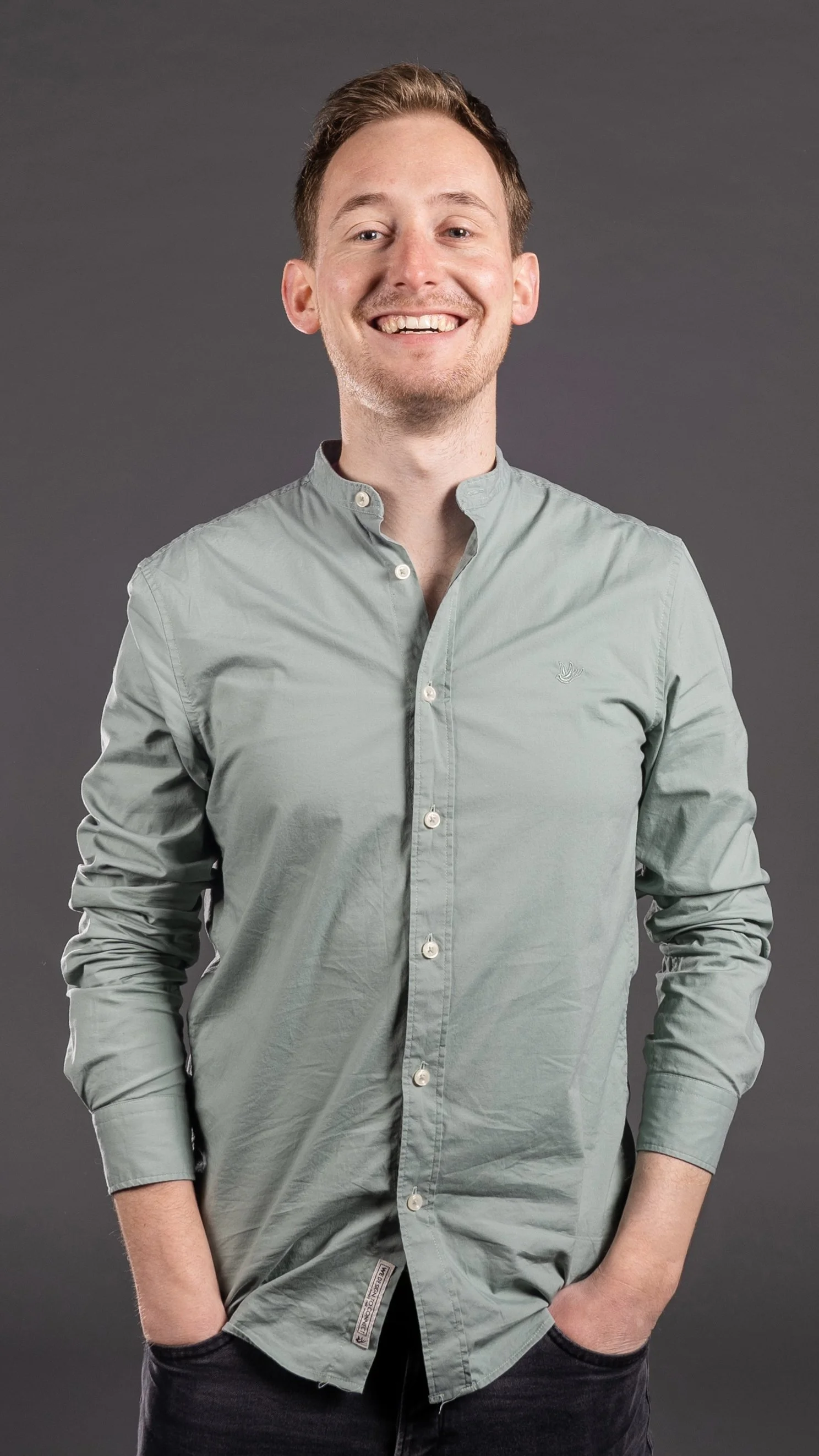 A smiling man with light brown hair and a light beard, wearing a light gray button-up shirt and black pants, standing against a gray background.