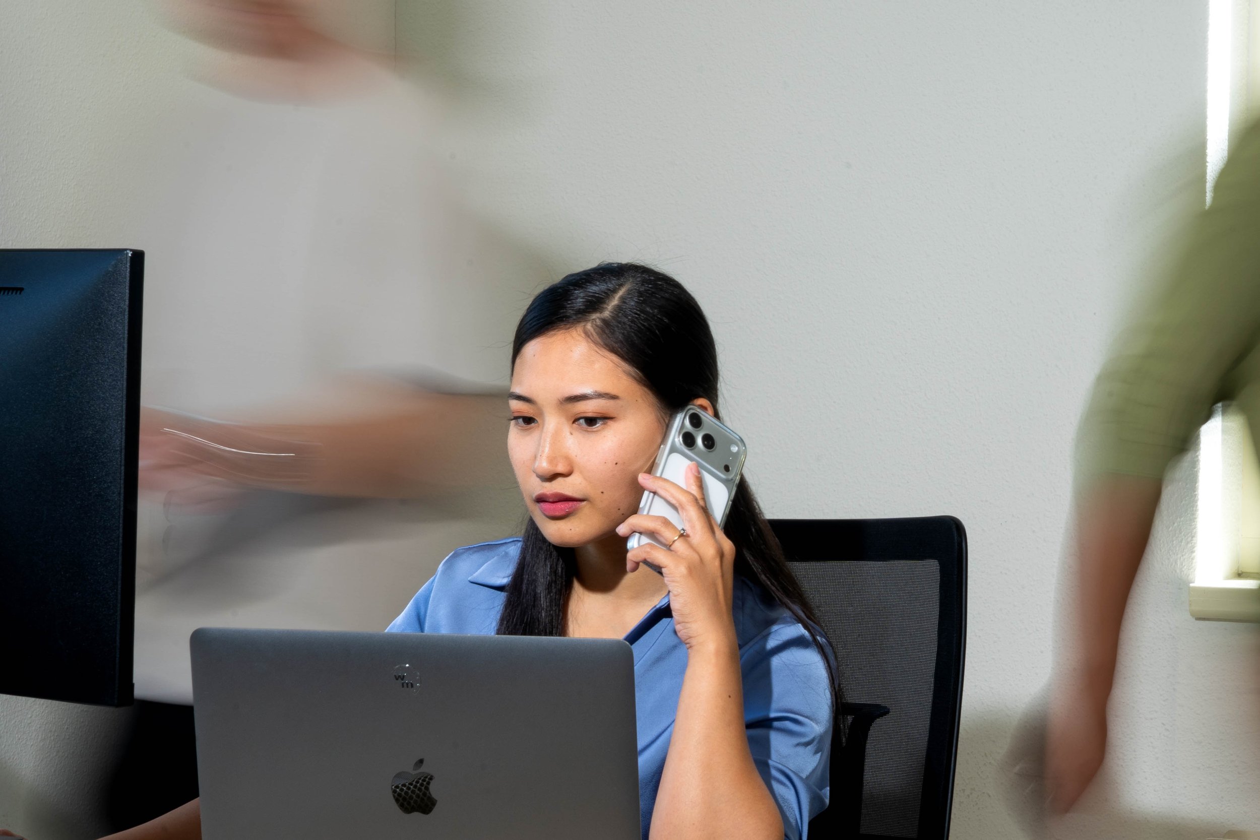 A young woman with long dark hair talking on her smartphone while working on a laptop in an office.