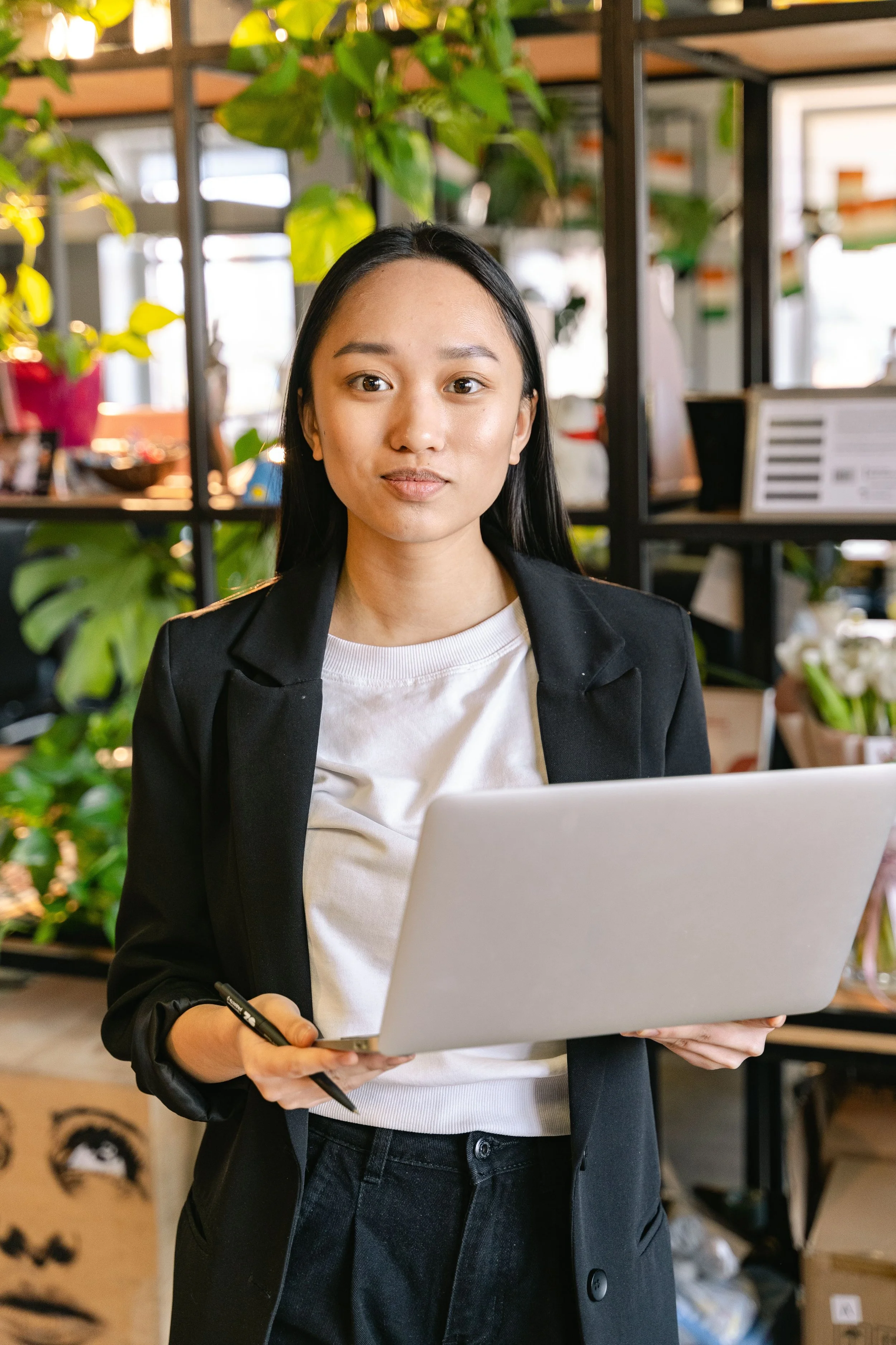 A woman with long black hair wearing a black blazer and white shirt, holding a laptop and a pen, standing in a room with plants and shelves in the background.