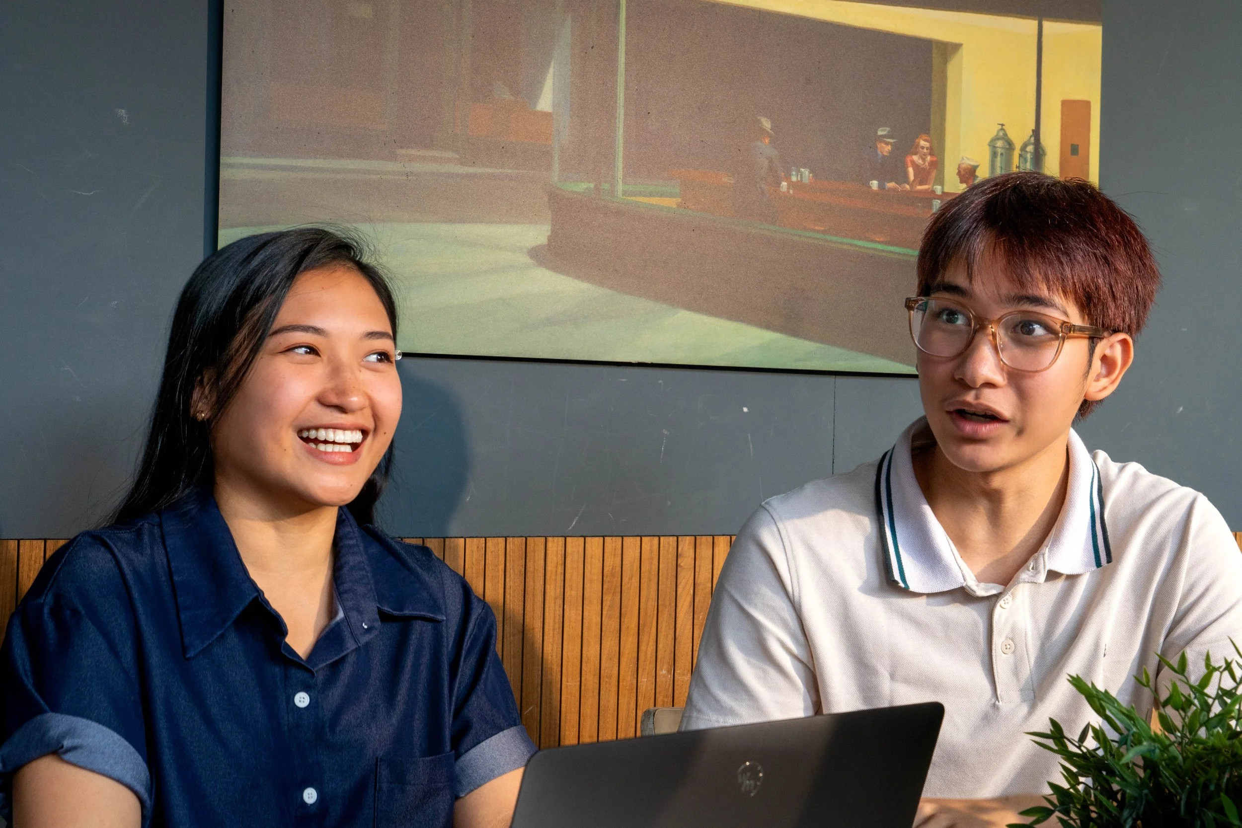 Two young people, a woman and a man, sitting at a table and looking at a laptop. The woman is smiling, and the man is speaking or explaining something. There is a projection of a restaurant scene on the wall behind them.