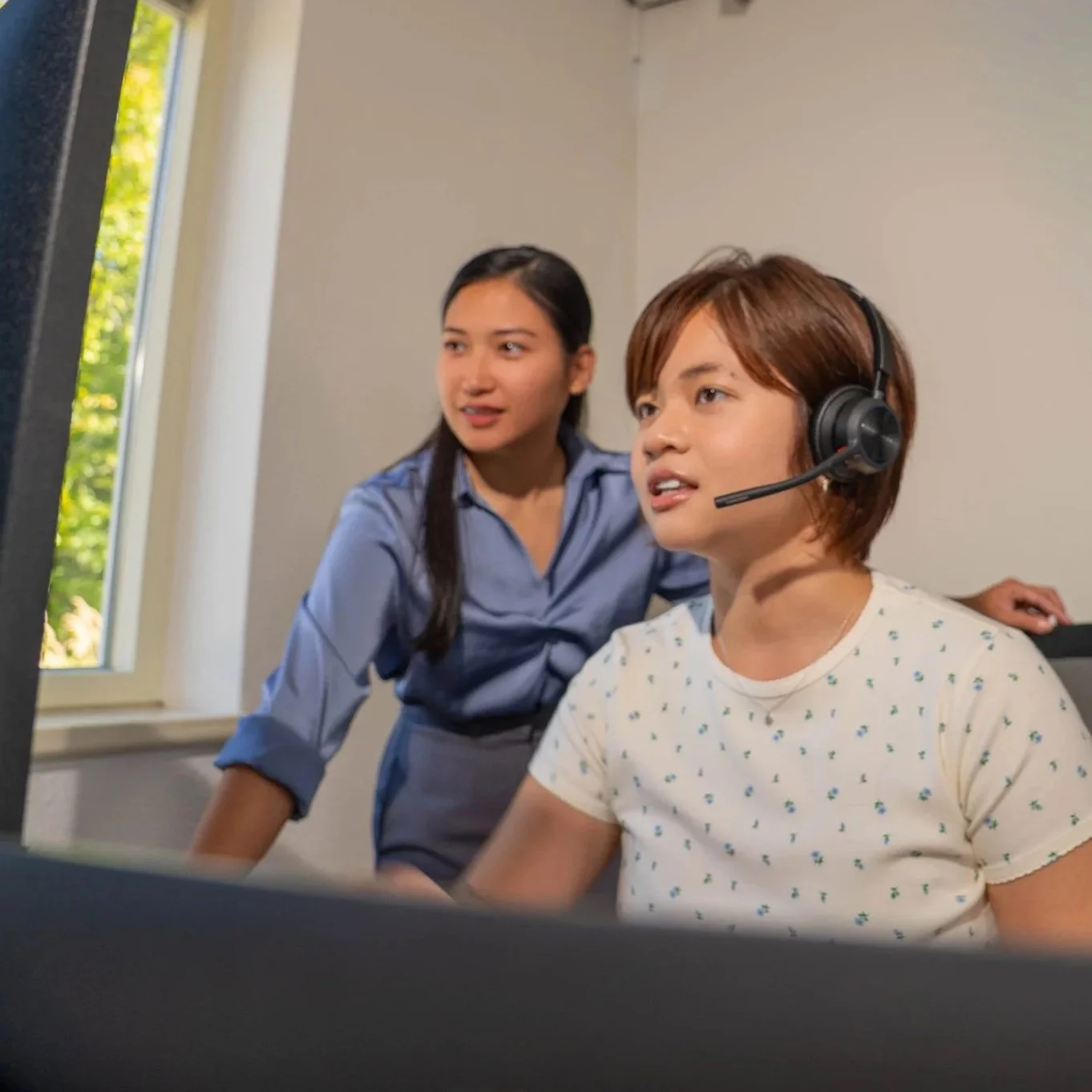 Close-up of smiling woman wearing a headset with microphone, line of similar women in background, all wearing headsets, in a call center or customer service environment.