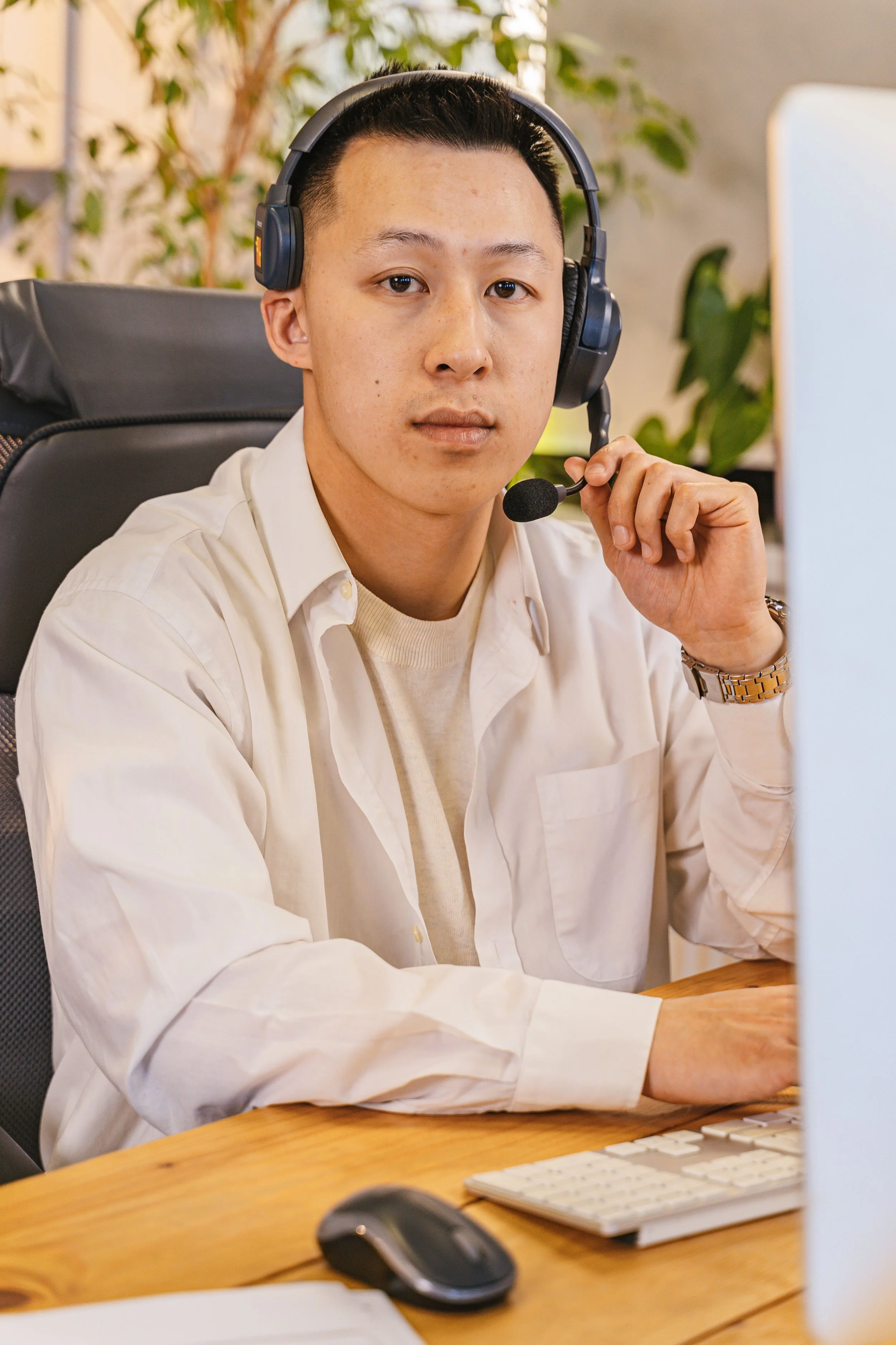A young man with light skin, short dark hair, and wearing a white shirt, sitting at a wooden desk with a computer, a computer mouse, and a keyboard. He is wearing a black headset with a microphone and looking directly at the camera. There are green plants and a beige wall in the background.