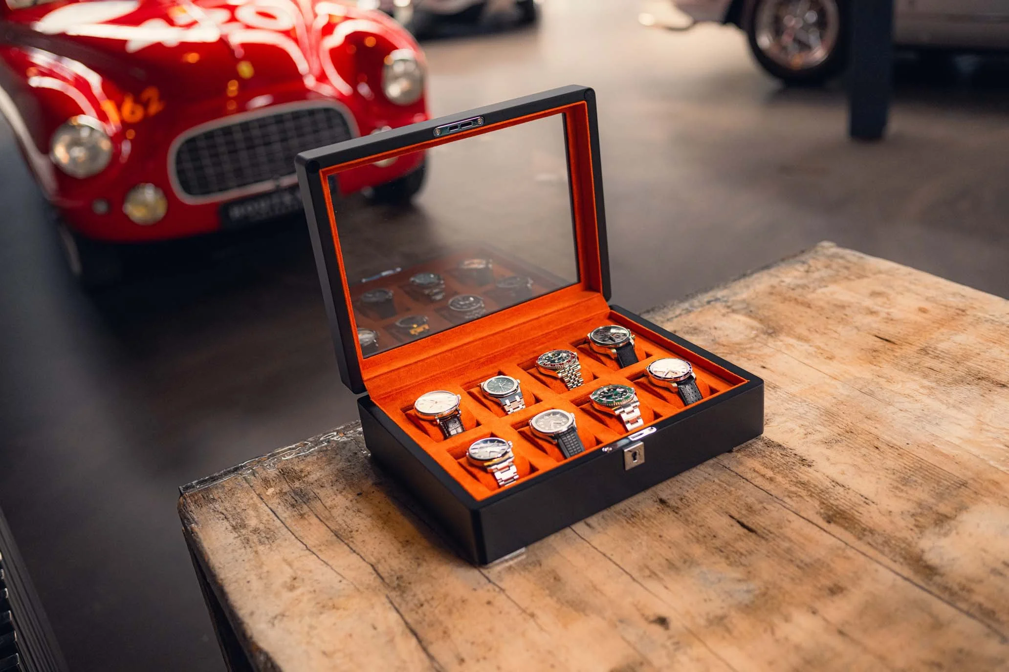 A black watch storage box with an orange interior, containing various wristwatches, is placed on a vintage wooden table. In the background, there is a red classic car and other vehicles in a garage or showroom.