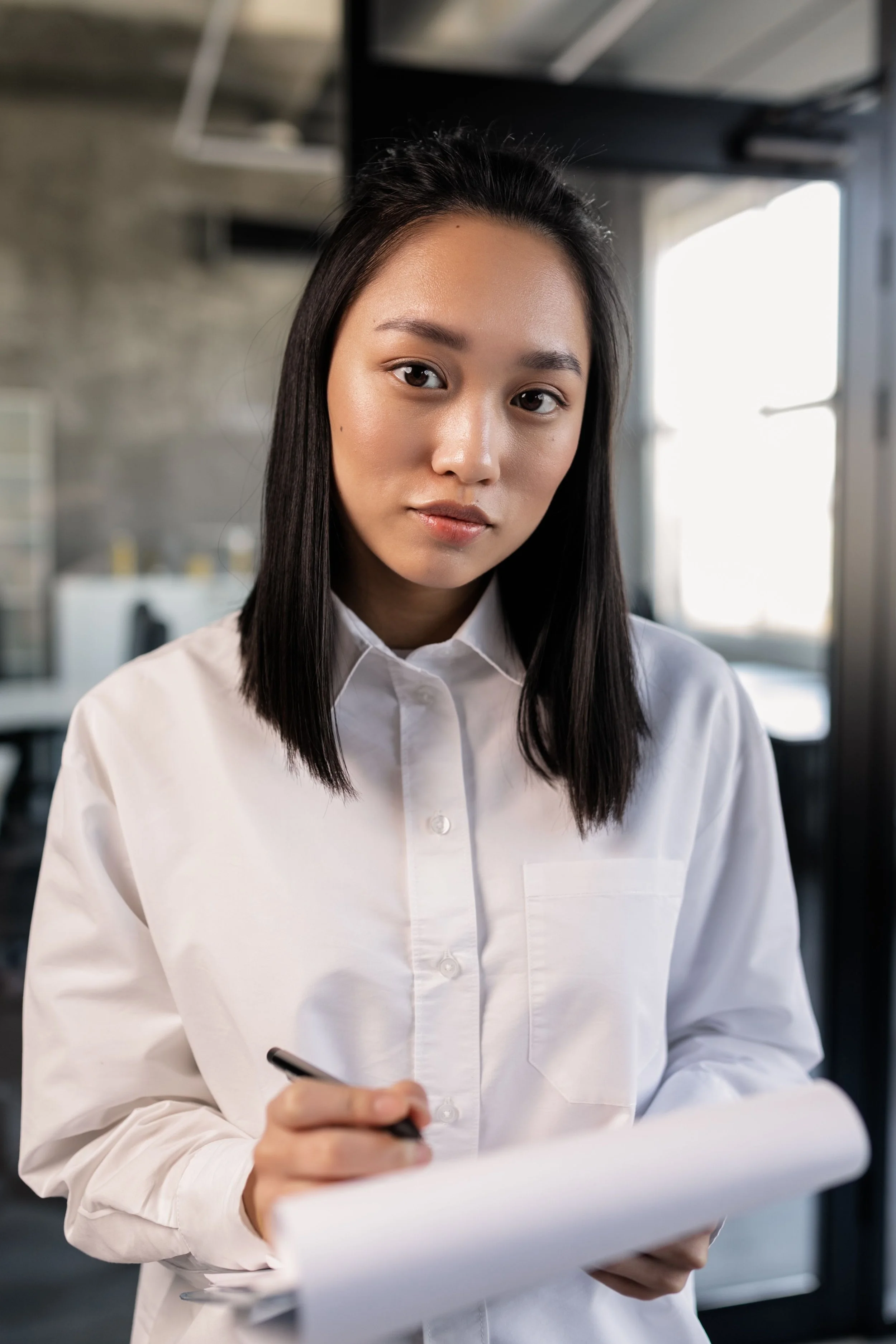 A young woman with straight black hair wearing a white button-up shirt, holding a pen, in an industrial or modern office setting near a window with natural light.
