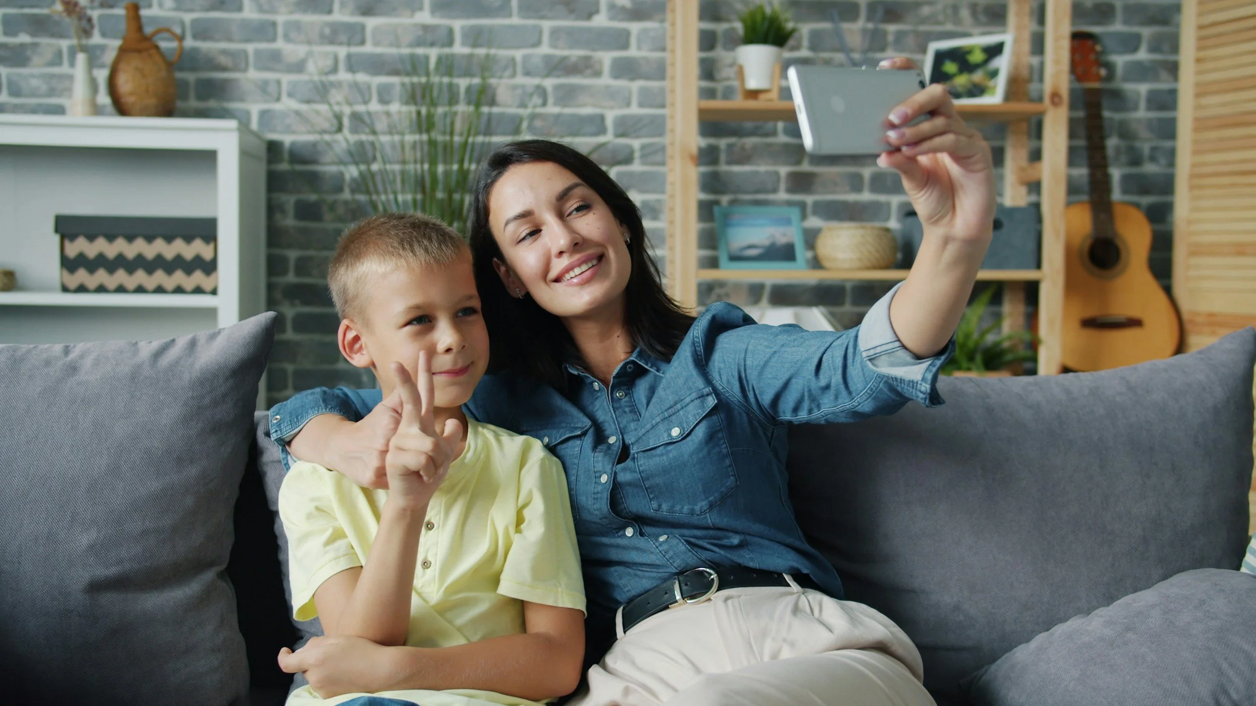 A woman and a young boy sitting on a gray couch taking a selfie together. The woman is smiling and holding a smartphone, while the boy is making a peace sign with his fingers and smiling. The background shows a decorative shelf, a guitar, and a brick wall.