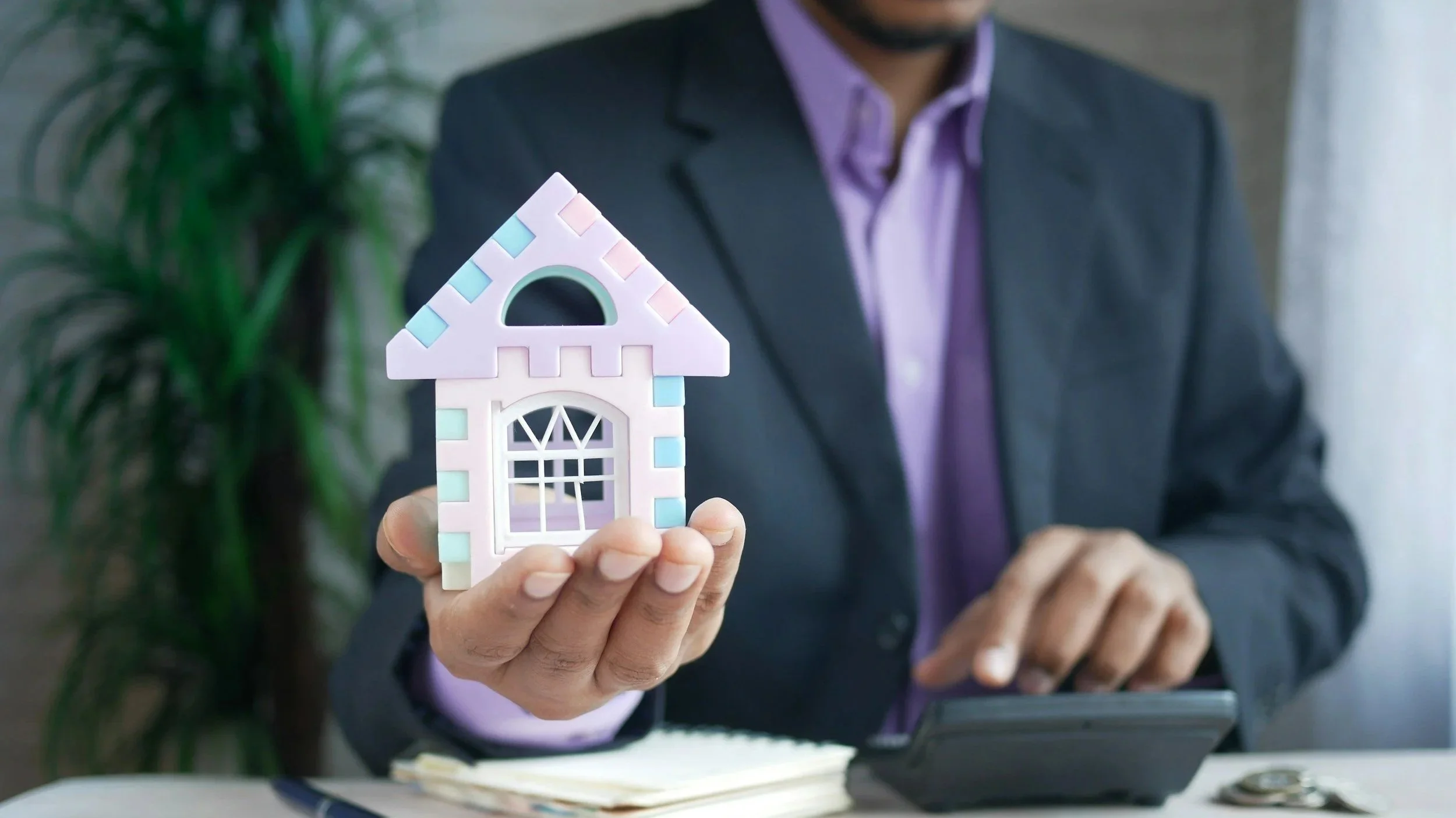 Person in a suit holding a small pink and blue house model during a real estate or financial discussion.