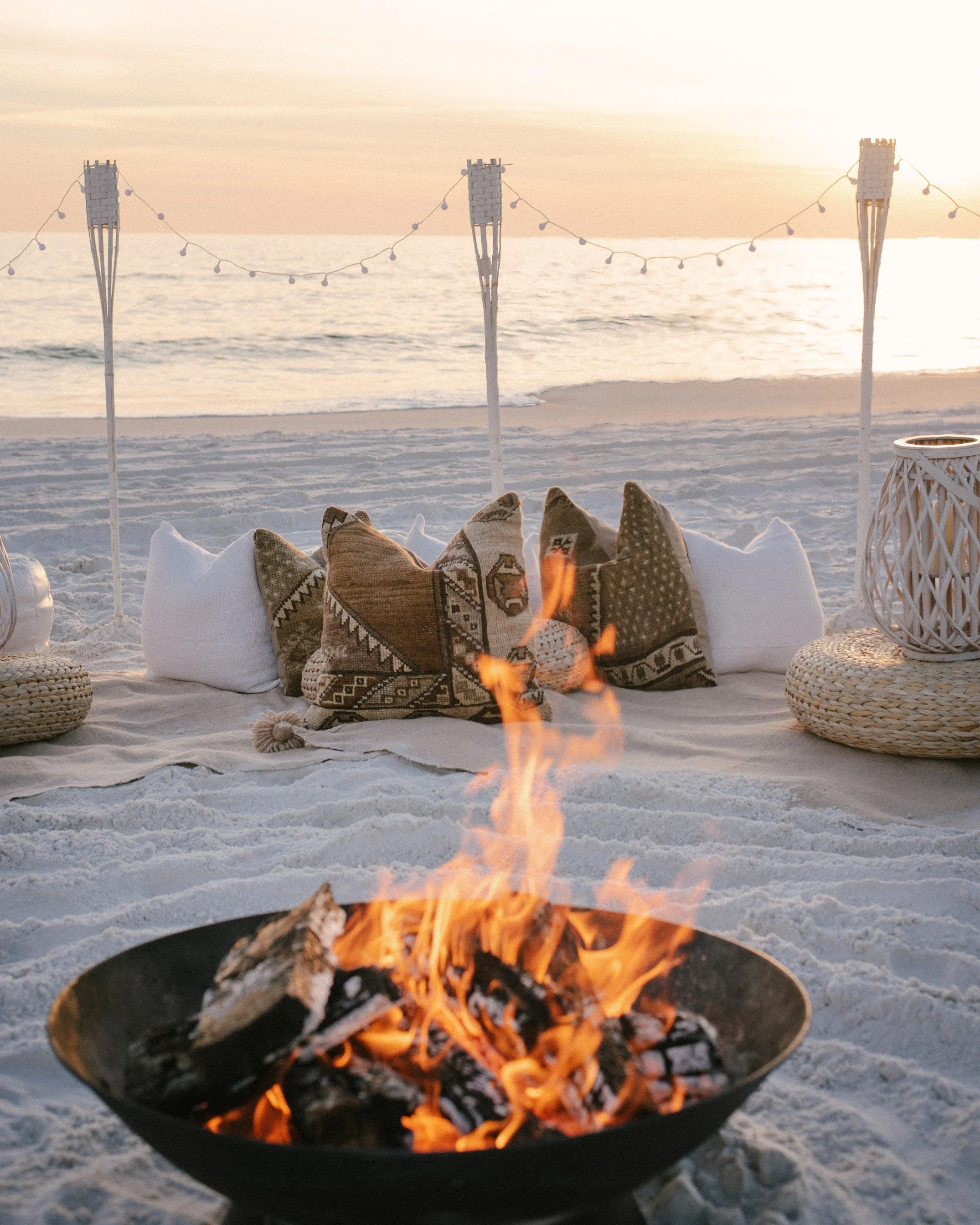 A close-up of a fire burning outdoors on the beach at sunset, with sand, decorative pillows, and lanterns nearby, and ocean and sky in the background.