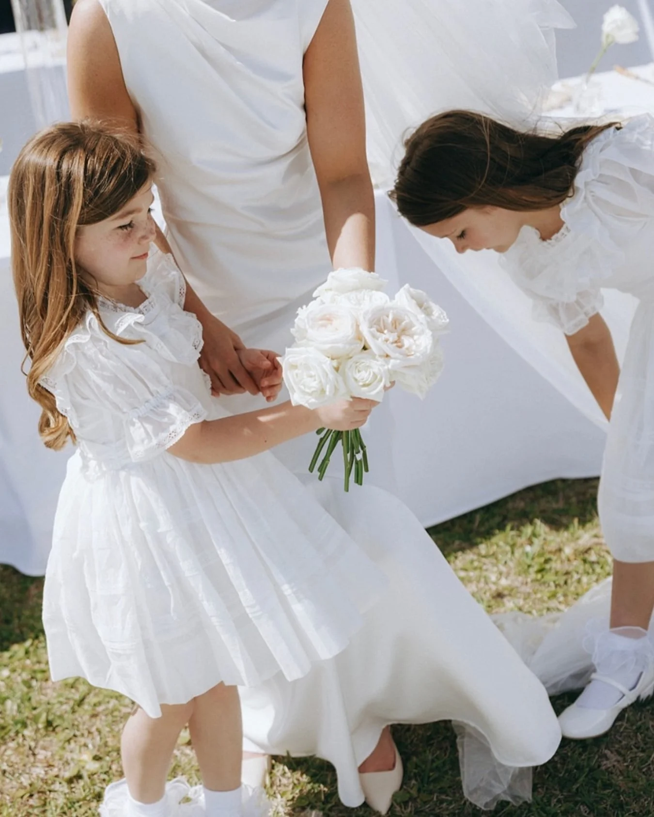 A young girl in a white dress holding a bouquet of white flowers at a wedding, with an older girl in a white dress leaning over to look at the flowers.