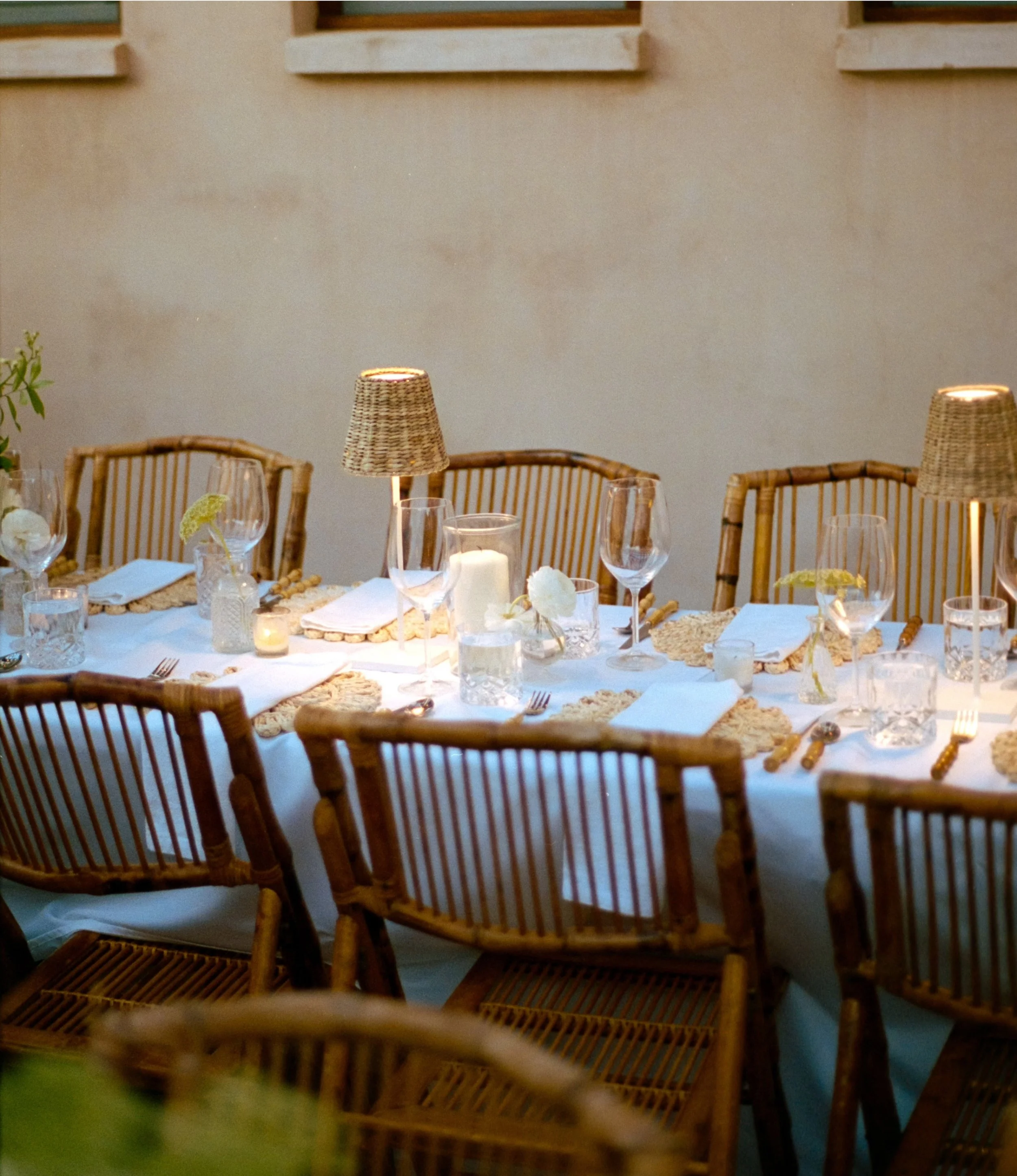 A dining table set with wine glasses, candles, and floral decorations, surrounded by wooden chairs, with wicker lamps hanging overhead in a warmly lit room.