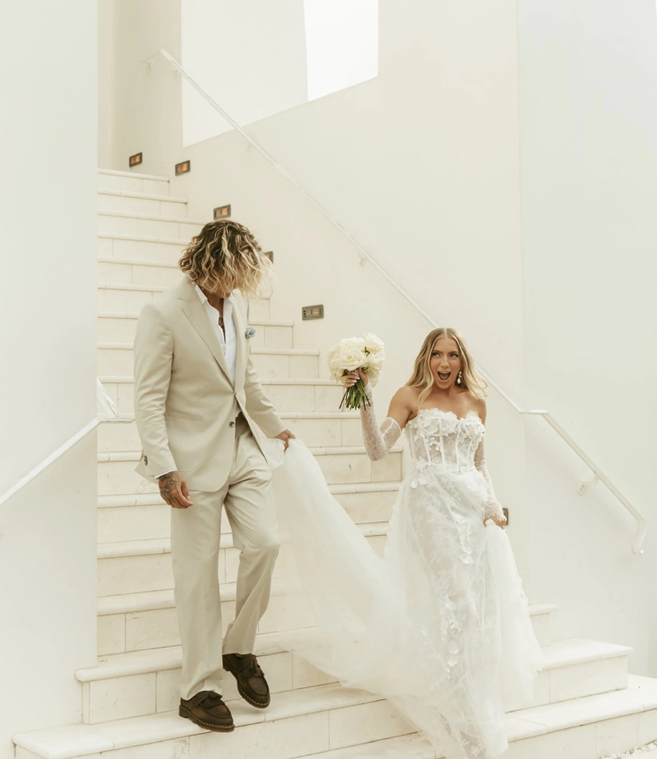 A couple is descending white marble stairs, dressed in wedding attire. The groom, in a light-colored suit and black boots, holds the bride's hand. The bride, in a lace wedding gown, is holding a bouquet of white roses and appears excited, with her mo