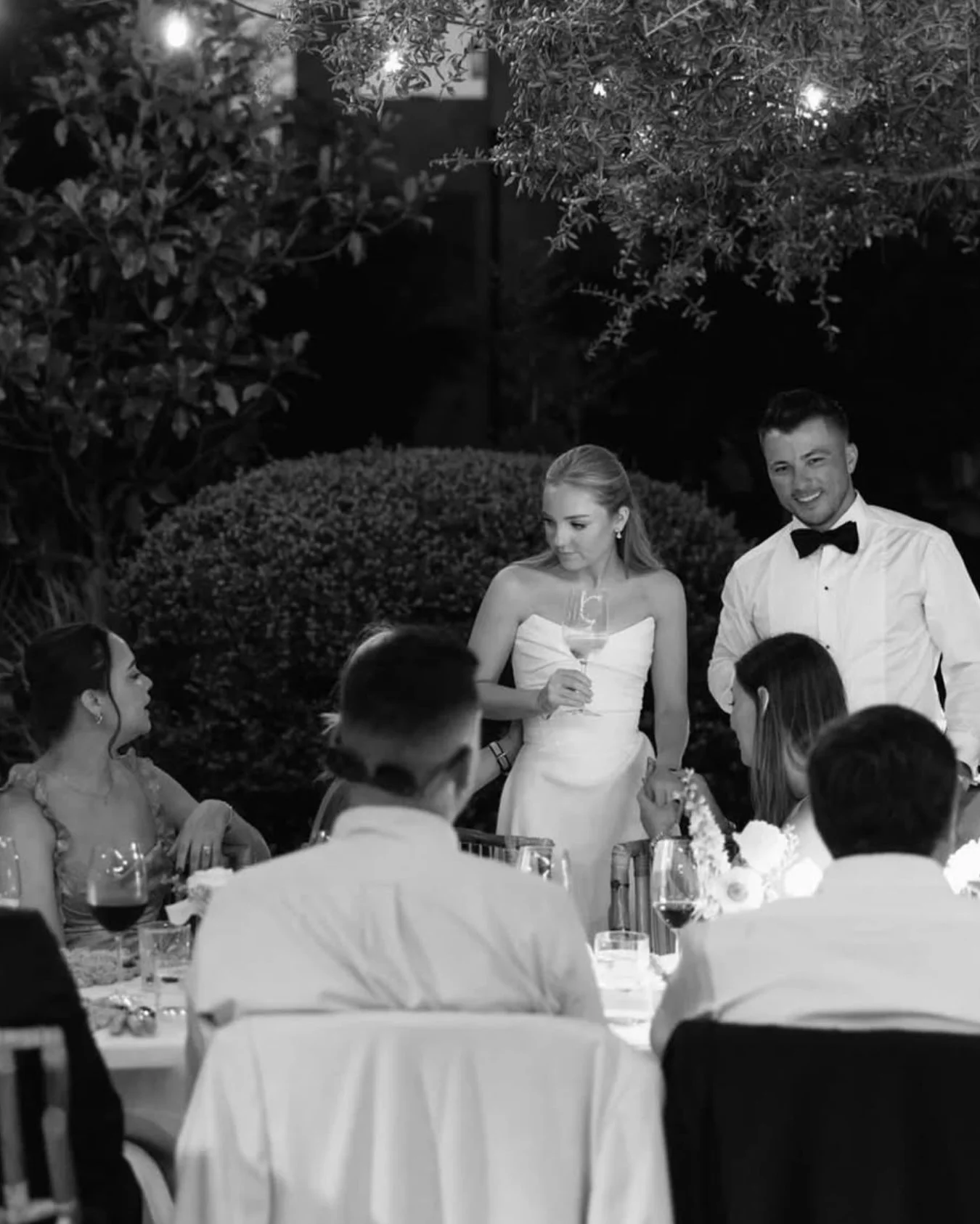 A black and white photo of a wedding reception dinner with guests seated at tables, a bride holding a glass, and a groom standing beside her wearing a tuxedo with a bow tie, outdoors at night with trees and lights in the background.