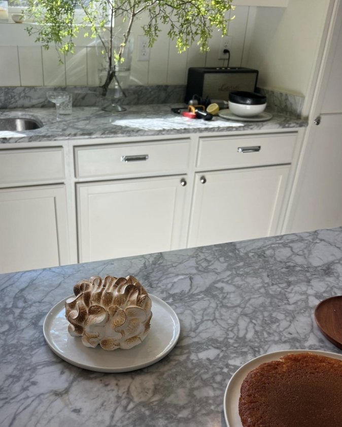 A kitchen with marble countertops and white cabinets. On the counter, there is a vase with green leafy branches, a glass, and a black and white bowl. On the foreground marble island, there is a meringue dessert on a white plate and a slice of cake on
