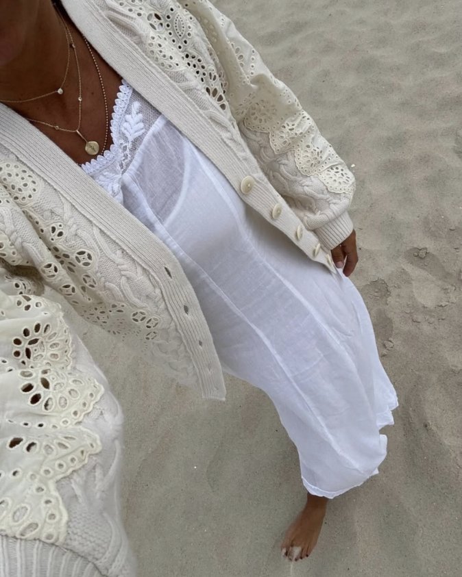Person wearing a white, lacy cardigan over a white dress, standing barefoot on sand at the beach.