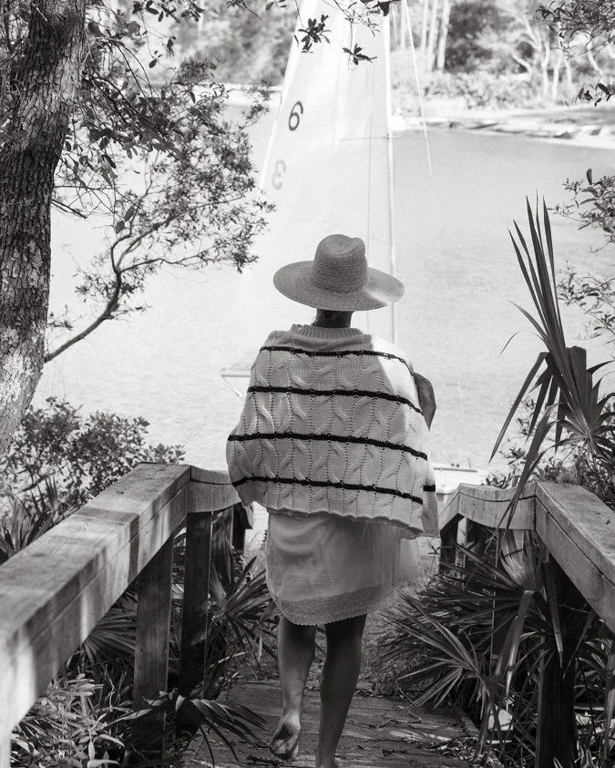 A person wearing a wide-brimmed hat and a shawl walking down a wooden path toward a body of water, surrounded by trees and plants.