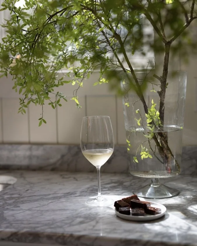 A glass of white wine, a plate of chocolates, and a large glass vase with water and green foliage on a marble countertop.