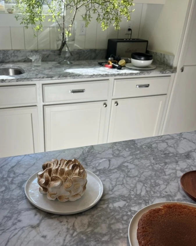 A kitchen with a marble countertop, a vase with green foliage, and a glass of water. On the countertop are some accessories, a bowl, and a small black device. In the foreground on the marble island are a cake with a swirl of icing and another cake on