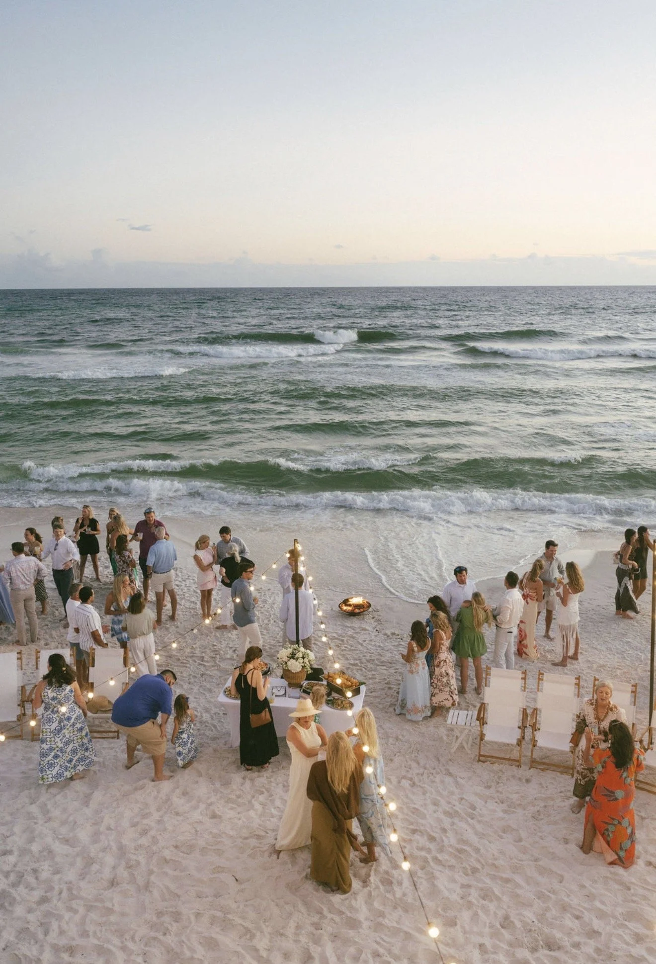 People gathering on a beach for a celebration or party during sunset, with ocean waves in the background and string lights decorating the scene.