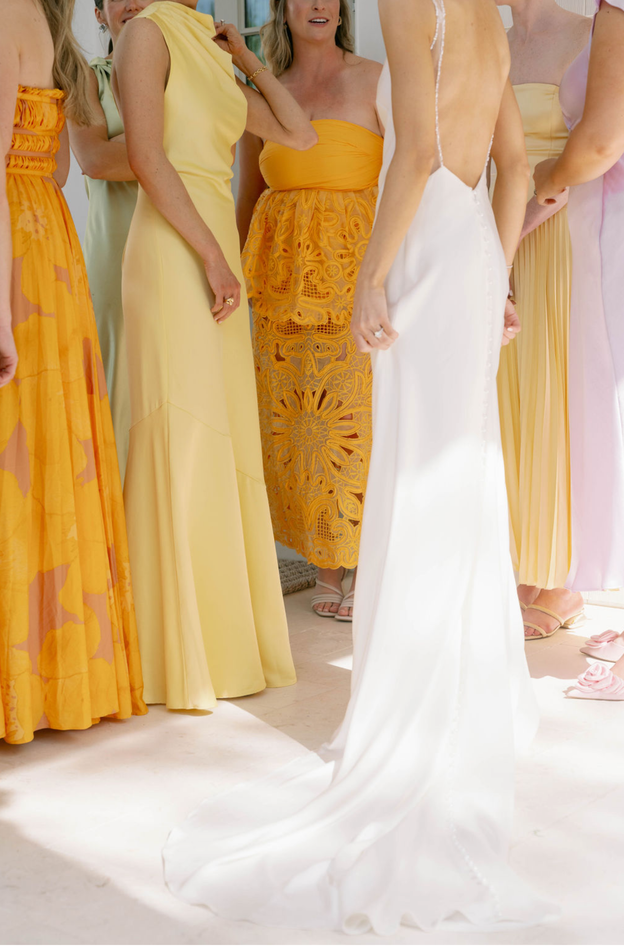 Women in colorful dresses, including a white wedding gown, standing indoors near a window during a wedding dress fitting or selection process.