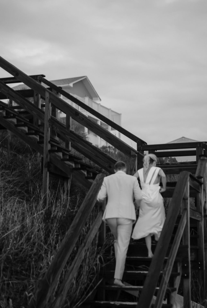 Couple walking up wooden stairs towards a house on a cloudy day, one man and one woman dressed in formal attire, with the woman in a long gown