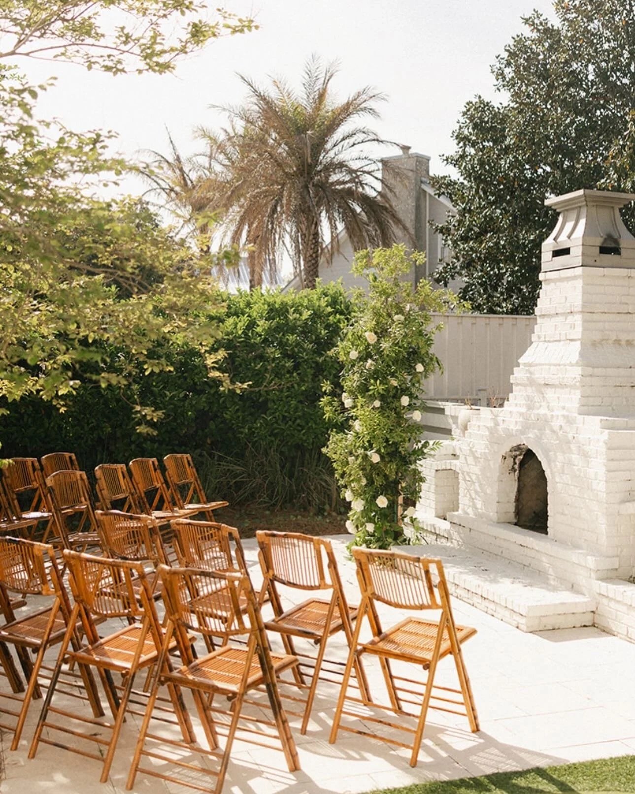 Outdoor gathering space with wooden chairs, a white brick fireplace, lush green bushes, and tall trees including a palm tree, with a house and fence in the background.