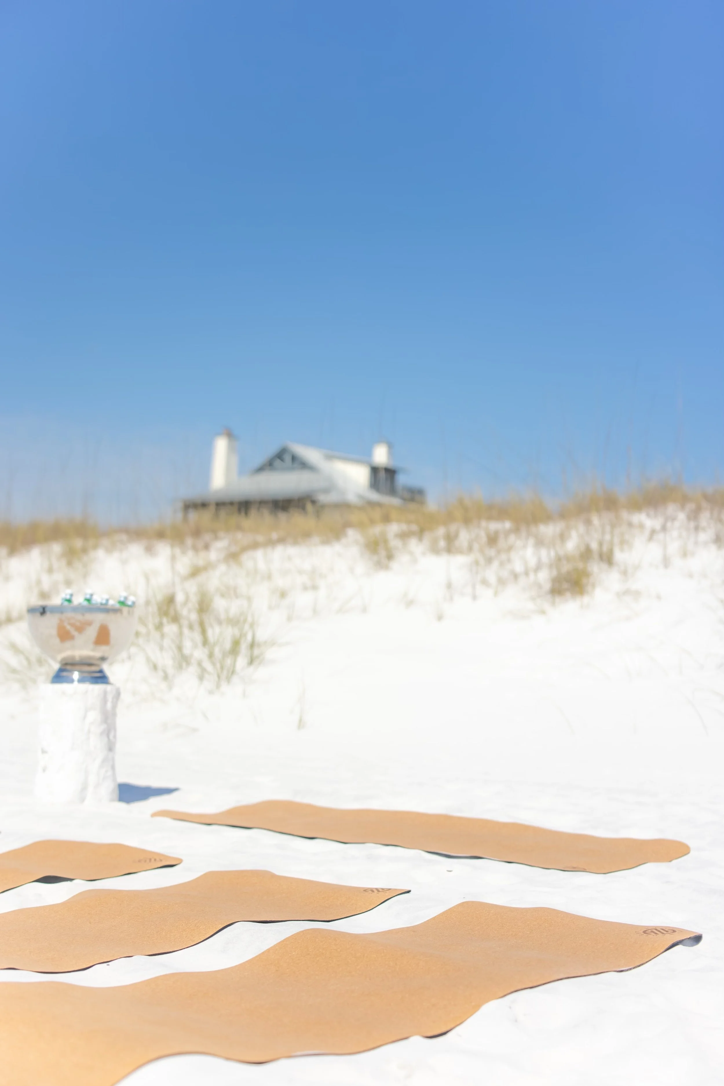 A beach scene with sand dunes, a house in the background, and a clear blue sky. There are brown yogamats laid out on the sand and an ice bucket with bottles on a post.
