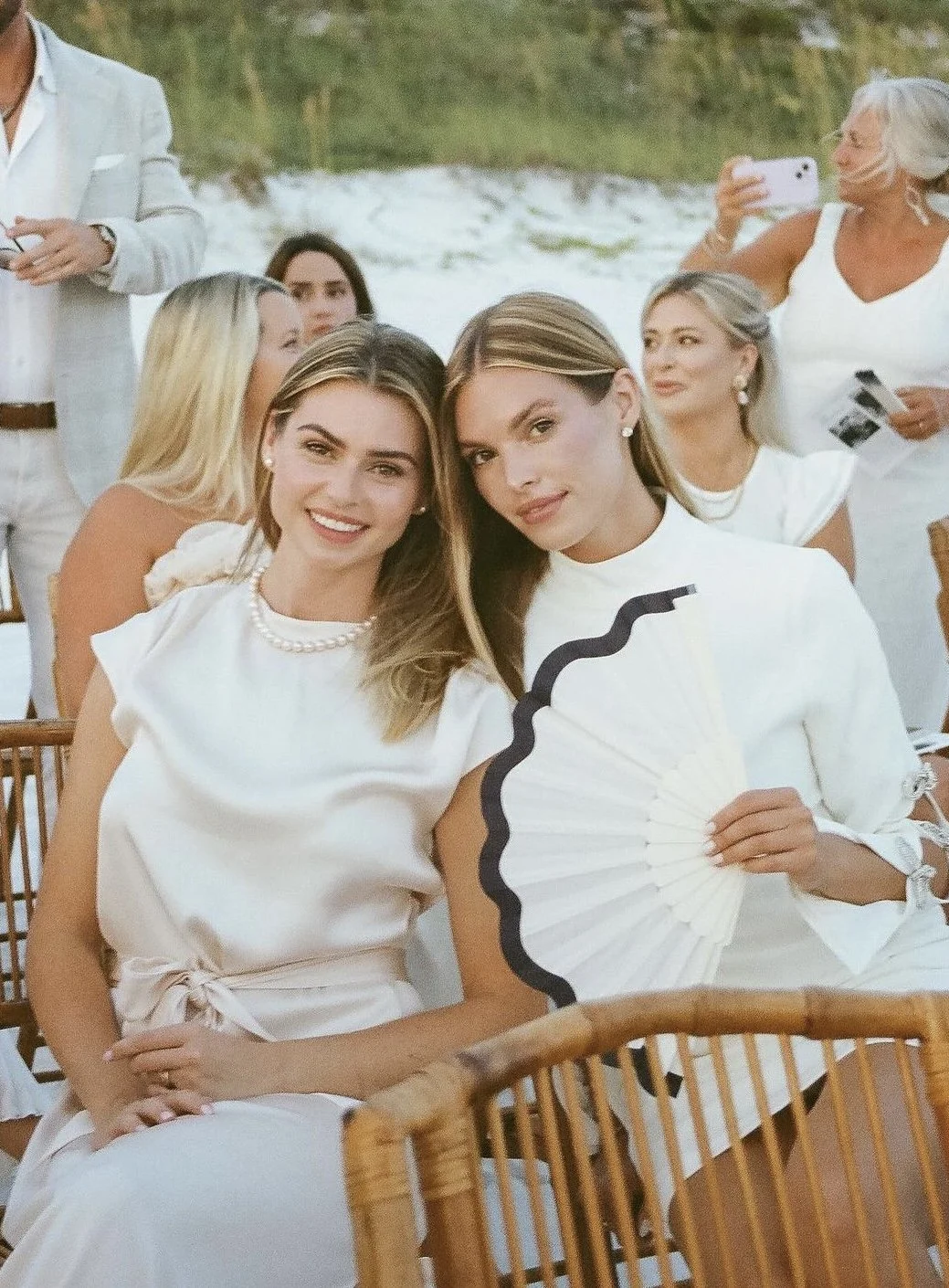 Two smiling women sitting together at an outdoor event, one holding a white fan with black trim, surrounded by other women in white attire, with a natural background of greenery and water.
